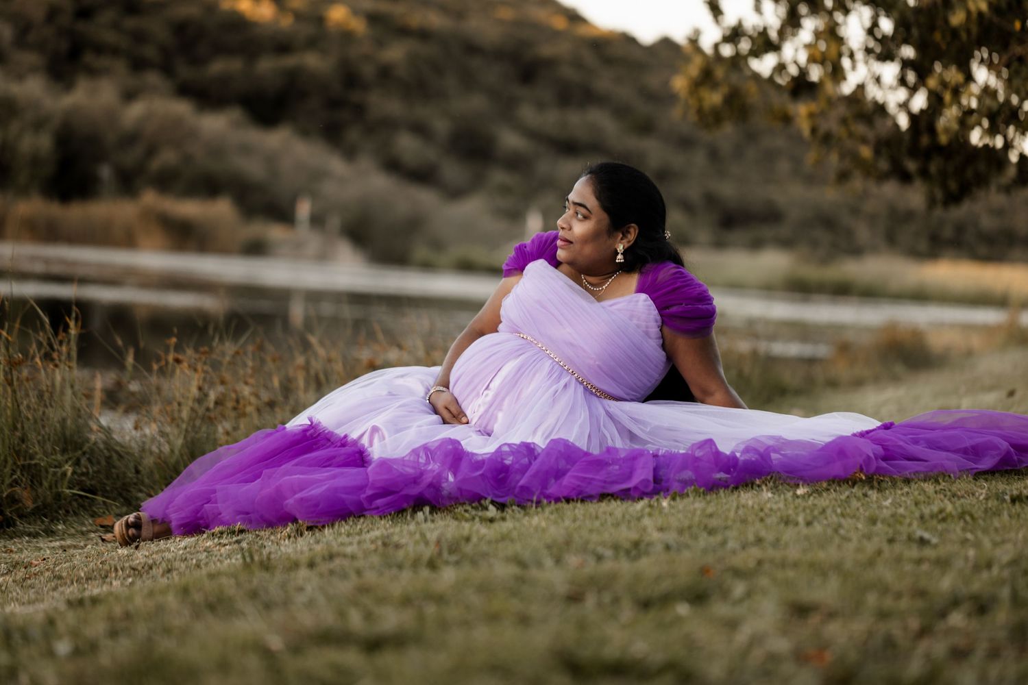 A person in a flowing white and purple dress lies gracefully on a grassy hillside during a sunset photoshoot.