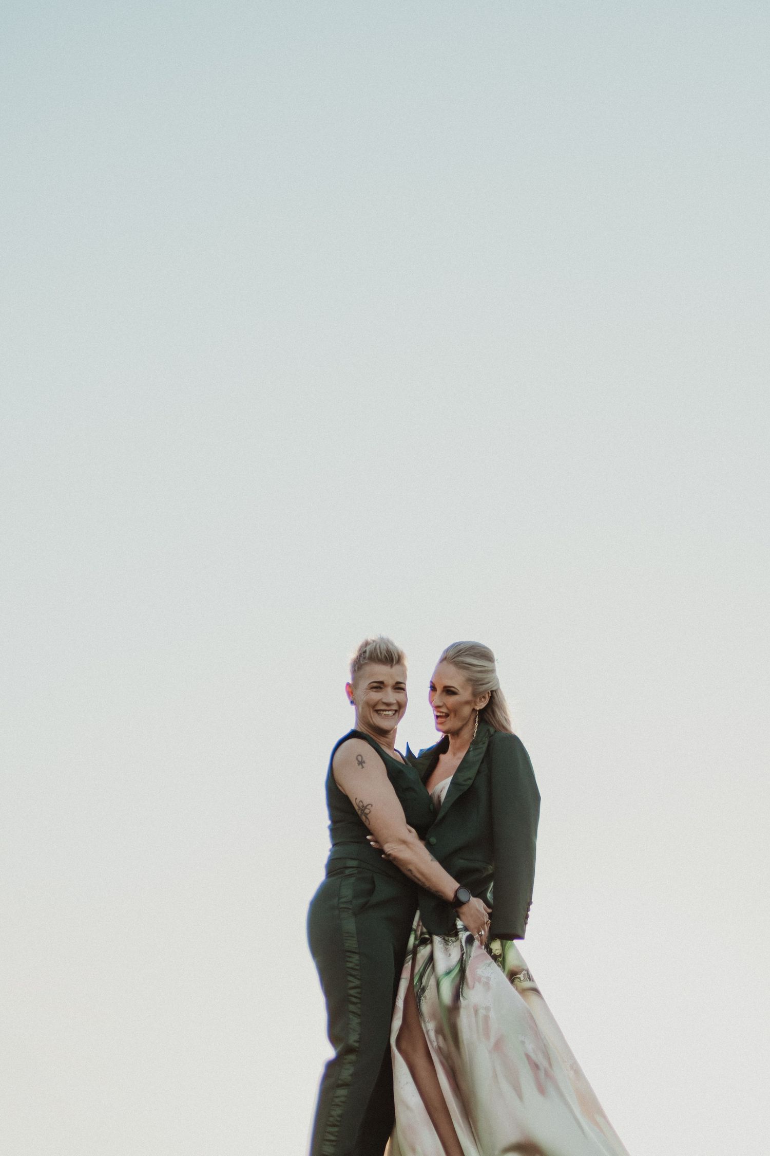 A couple embraces against a light sky background, one wearing a dark vest and the other in a flowing dress.