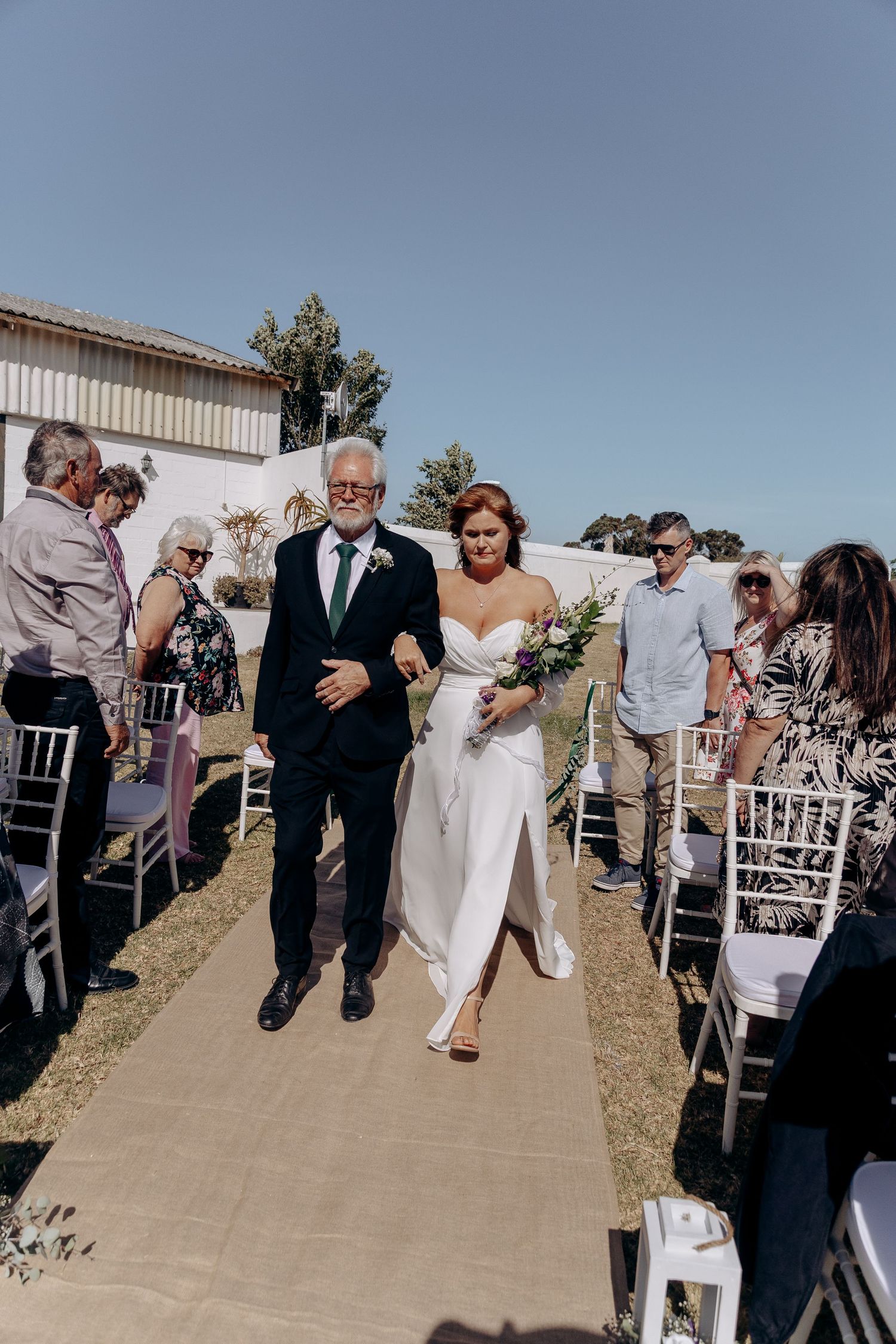 Bride walks down outdoor aisle with older gentleman, holding bouquet, guests seated on white chairs under clear blue sky.
