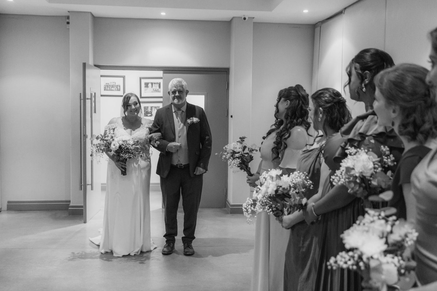 Black and white wedding photo of bride walking with escort past bridesmaids holding bouquets in elegant venue.