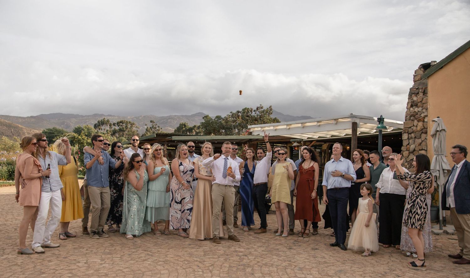 Large wedding party group photo outdoors at rustic venue with mountain backdrop and overcast sky.