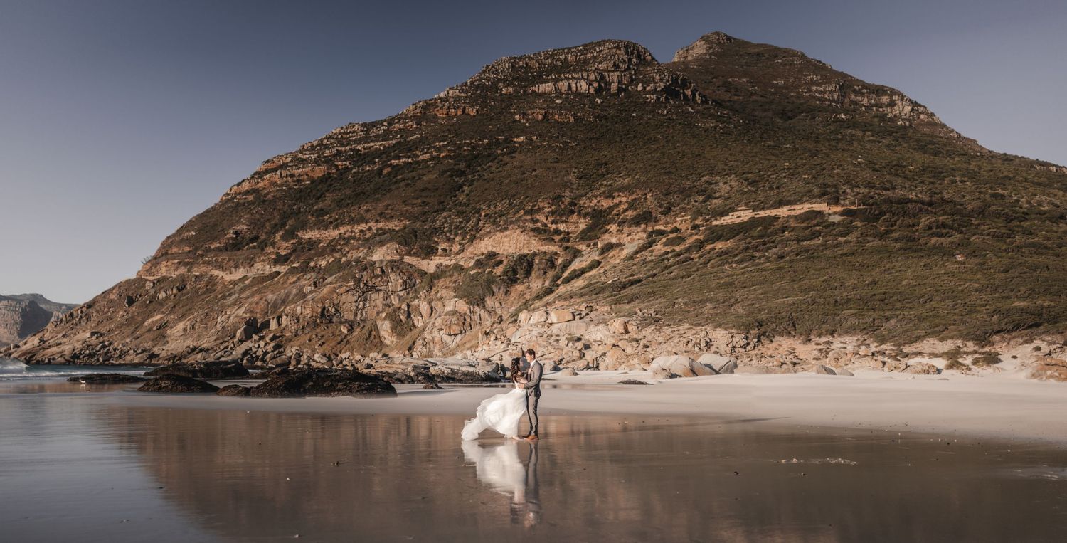 Couple in wedding attire standing on a reflective beach with dramatic rocky mountain backdrop.