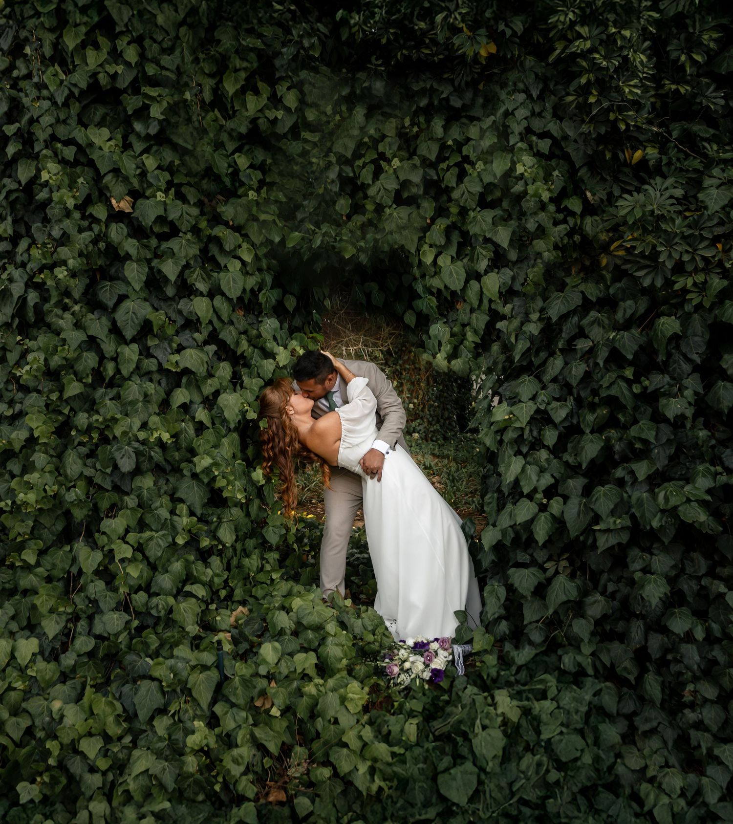 Couple sharing a romantic kiss in a lush green ivy-covered archway, bride in white gown holding purple flowers.
