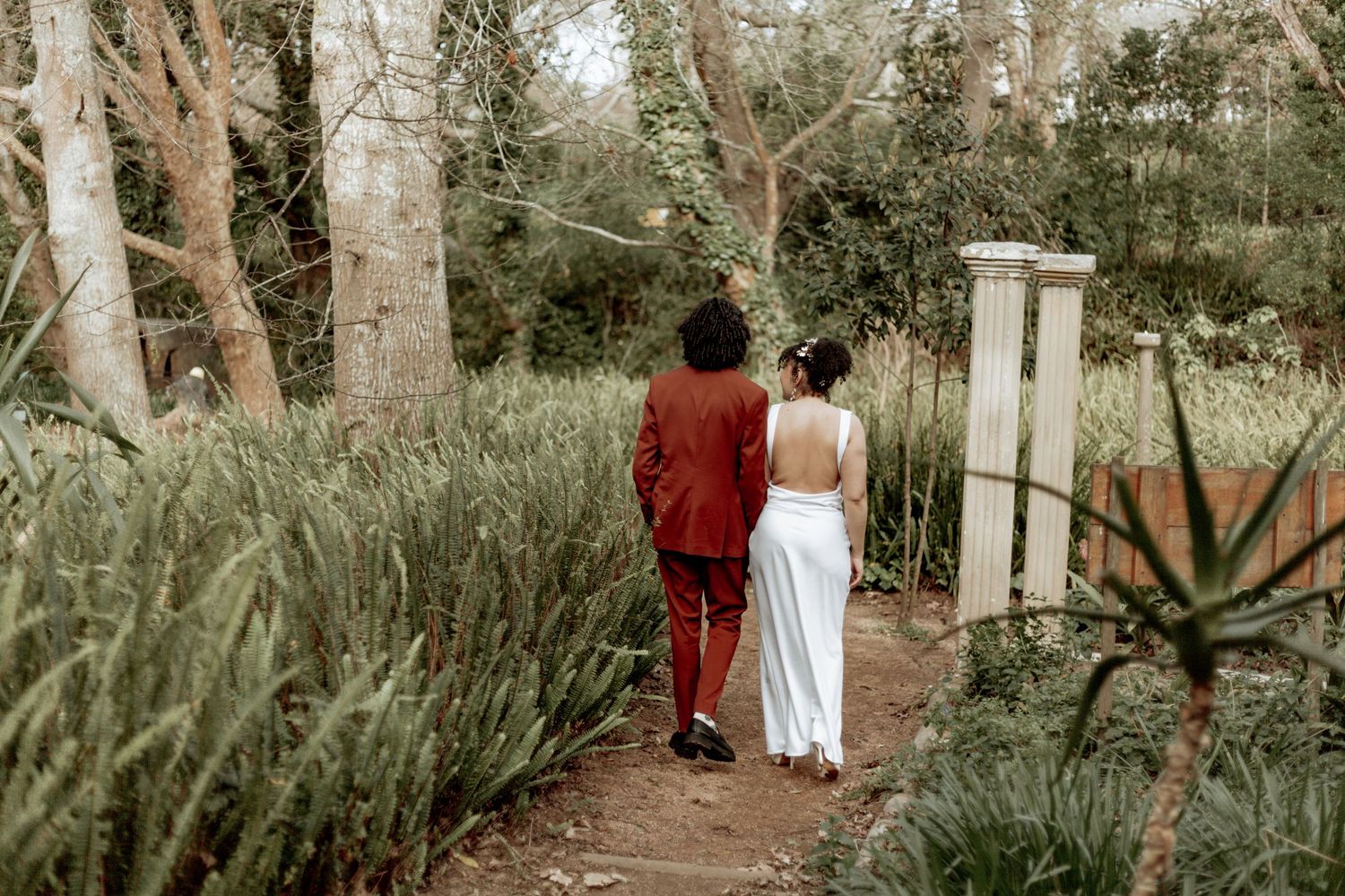 Two people walk down a lush garden path lined with tall trees and tropical plants in an outdoor setting.