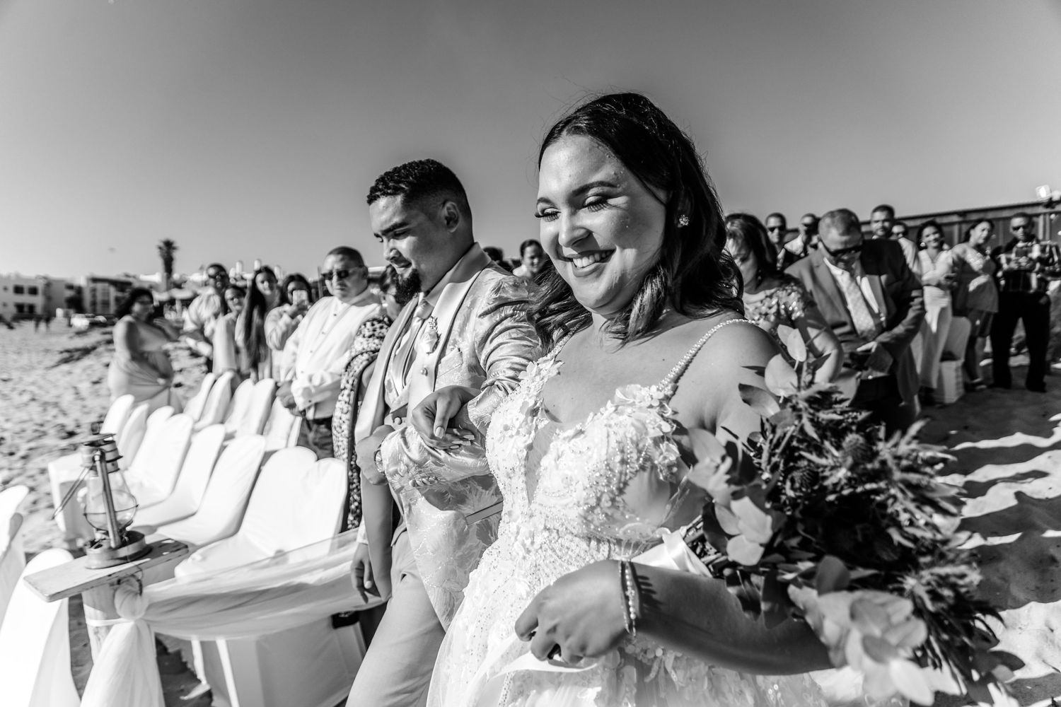 Black and white photograph of a couple during an outdoor beach wedding ceremony smiling and holding hands.