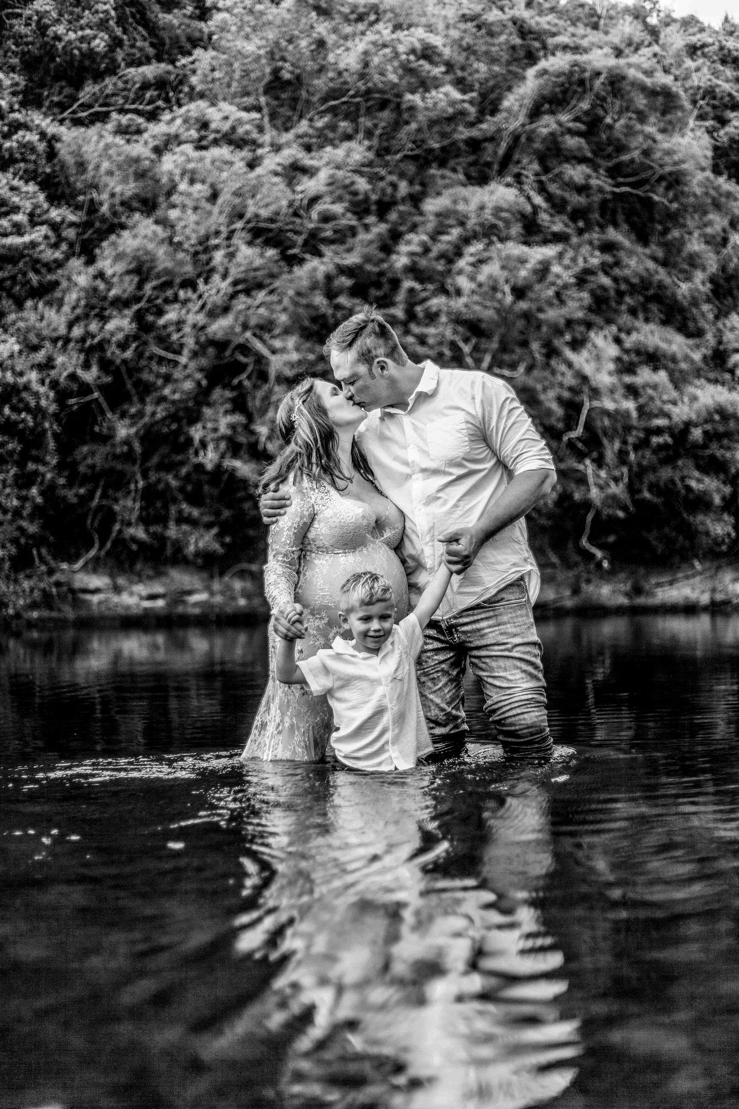 A tender family moment captured in black and white as they stand together in shallow river water surrounded by trees.