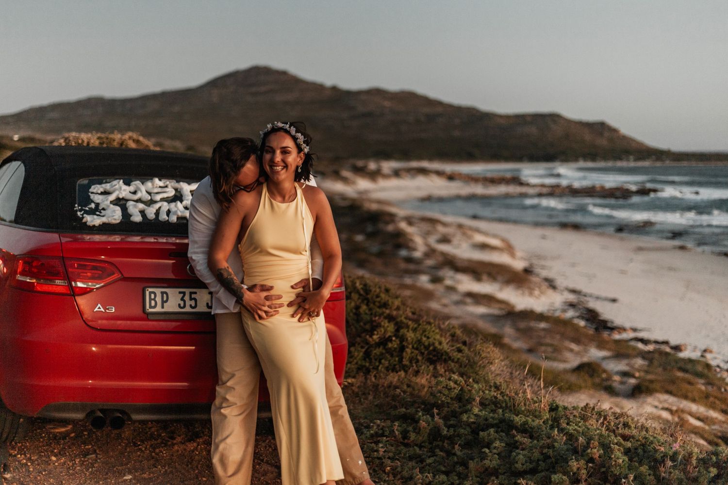 A couple poses in casual attire near the ocean coastline and red car during golden hour at a scenic beach location.