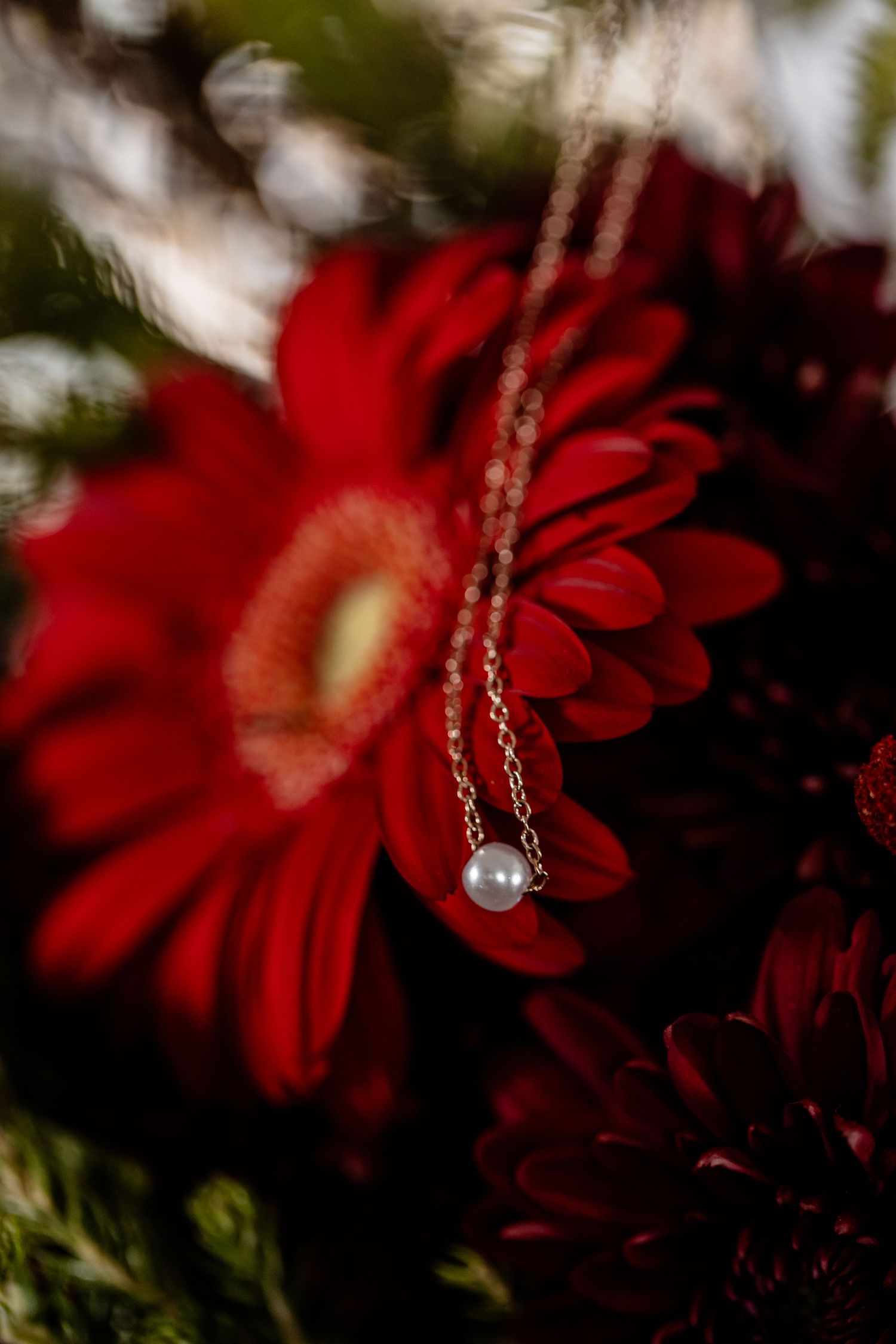 A delicate silver necklace with a pearl charm hangs in front of vibrant red flowers against a dark blurred background.