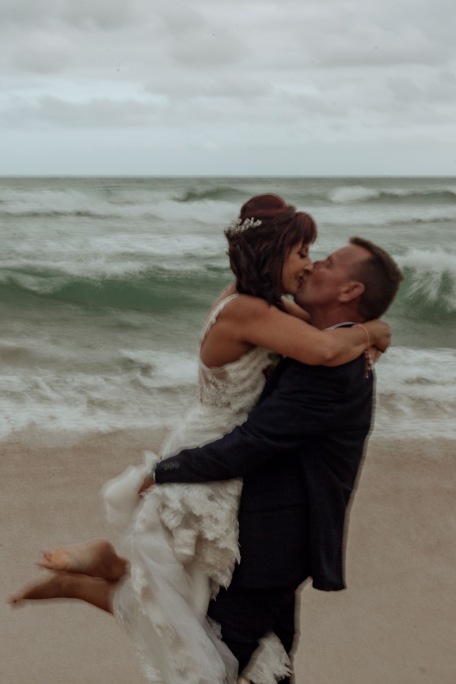 A romantic beach wedding moment as couple embraces on sandy shore with crashing waves in the background.