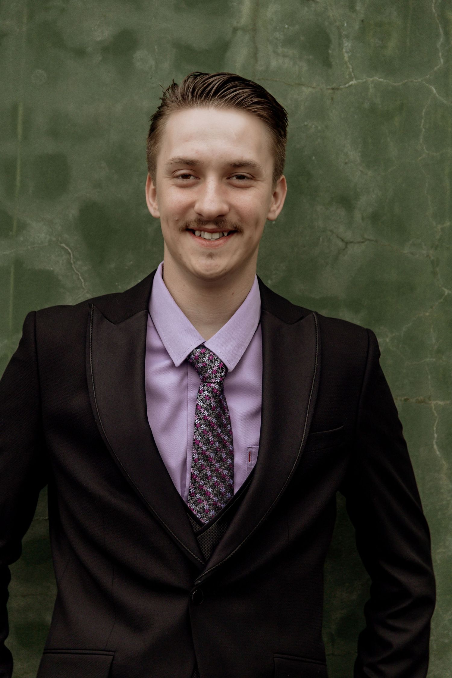 A person in a black suit and lavender dress shirt with patterned tie photographed in three similar poses against green wall.