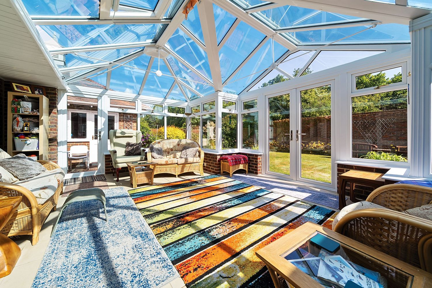 Bright sunroom interior with glass ceiling, colorful striped area rug, and wicker furniture overlooking backyard garden.