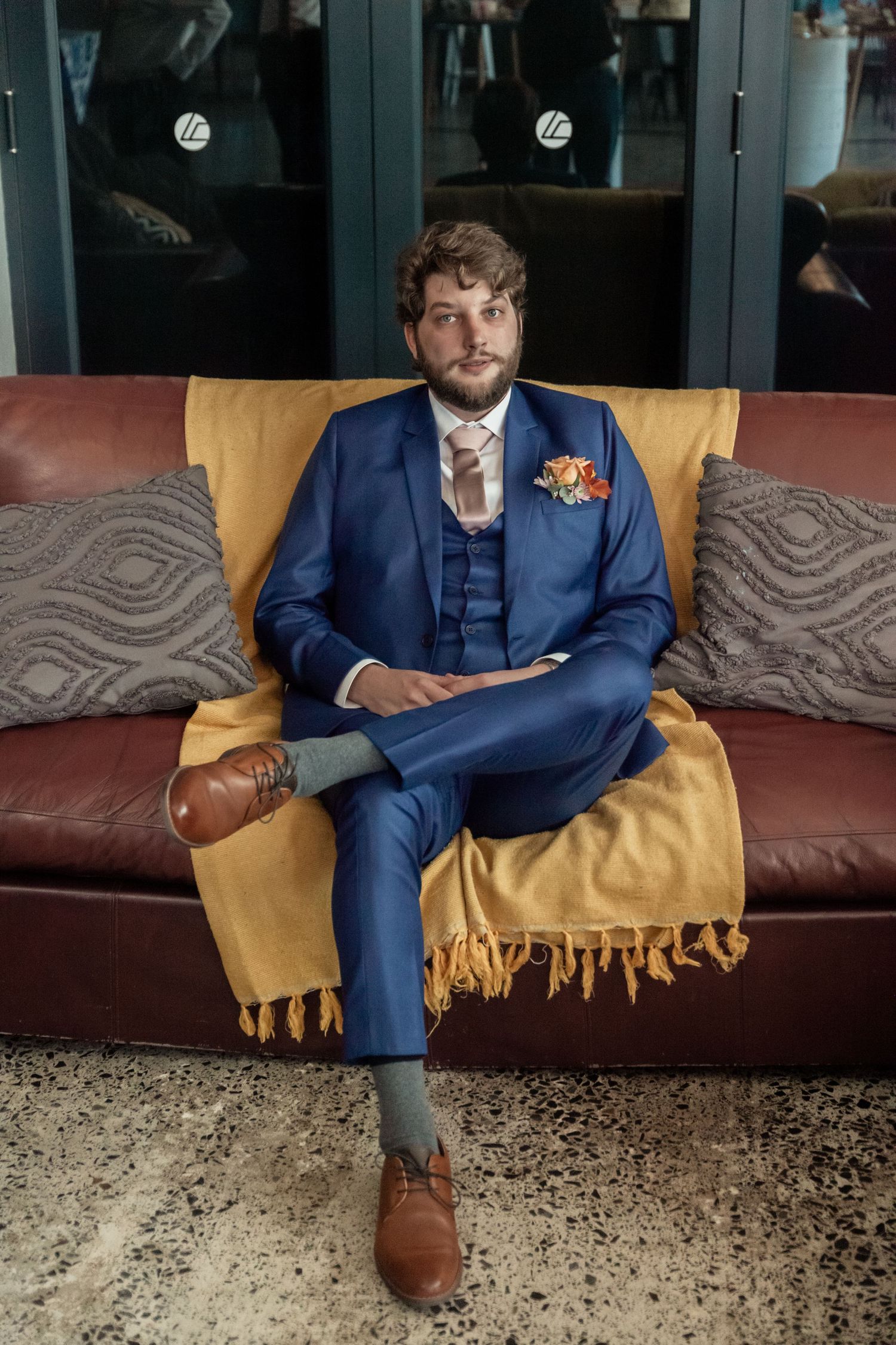Person in navy blue suit sits on yellow couch showing different poses and angles of their outfit and brown shoes.
