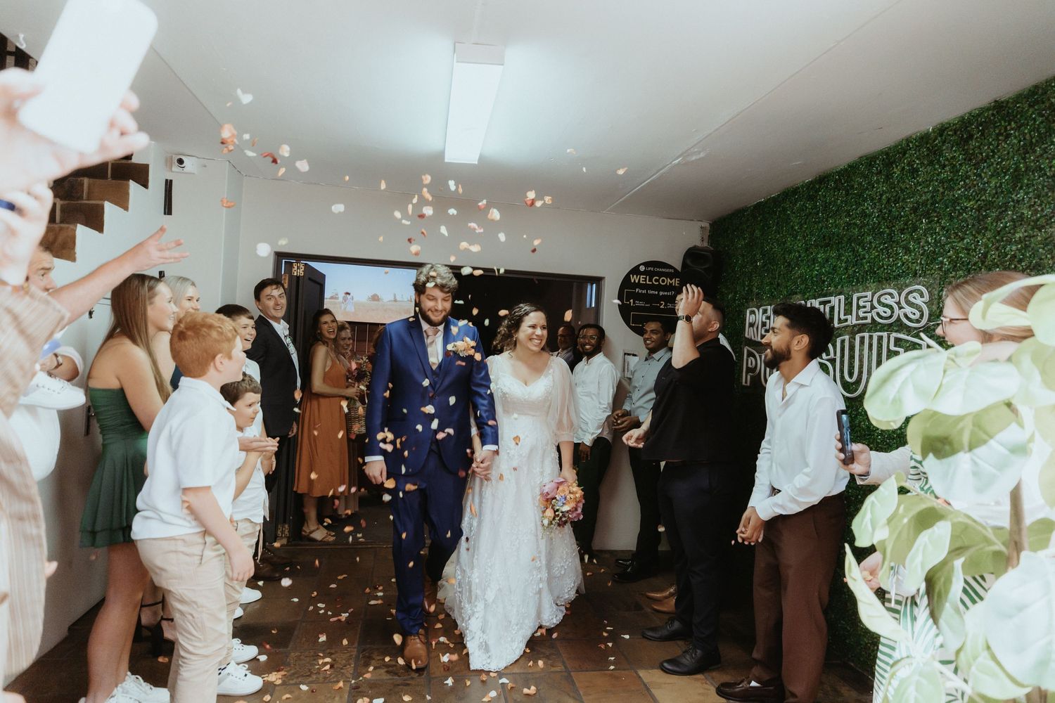 Wedding guests dance and celebrate on a white dance floor under twinkling lights at an indoor reception venue.