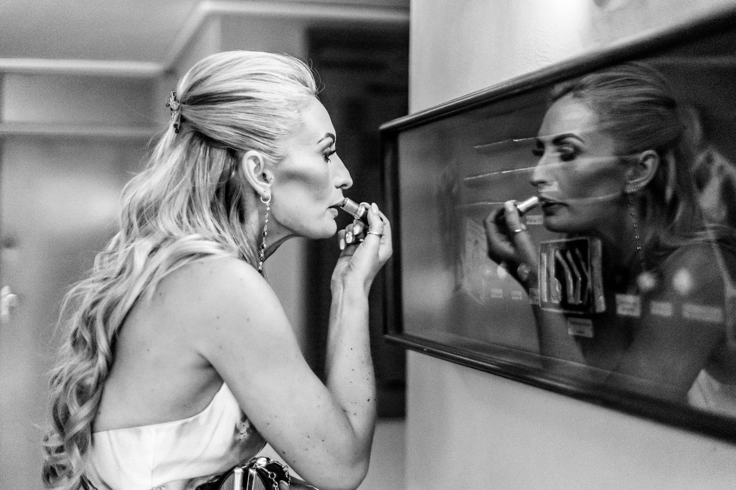 A woman with long blonde hair applies lipstick while looking at her reflection in a mirror, captured in black and white.