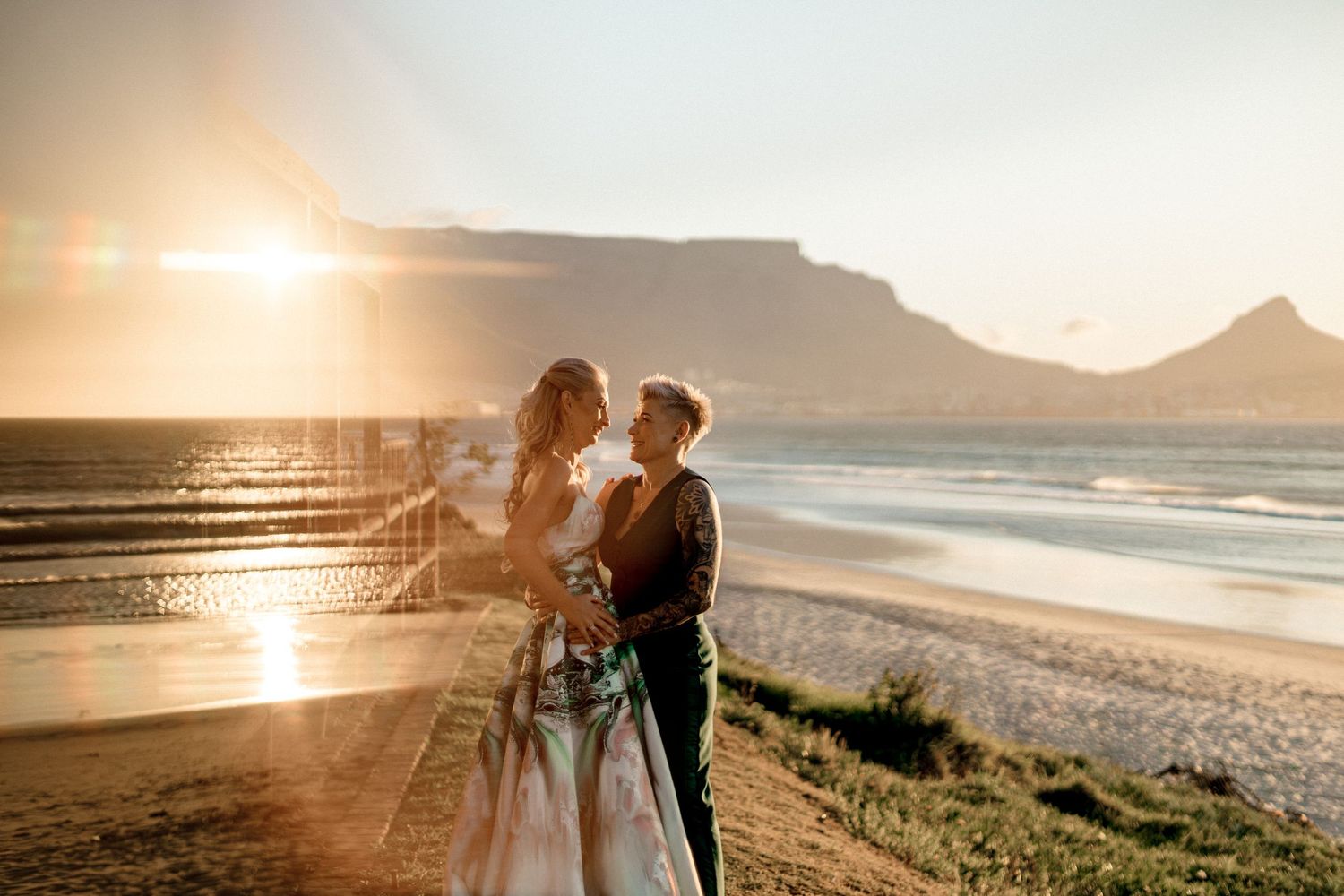 Couple embraces on scenic beach at sunset with golden light reflecting off ocean and mountains in background.