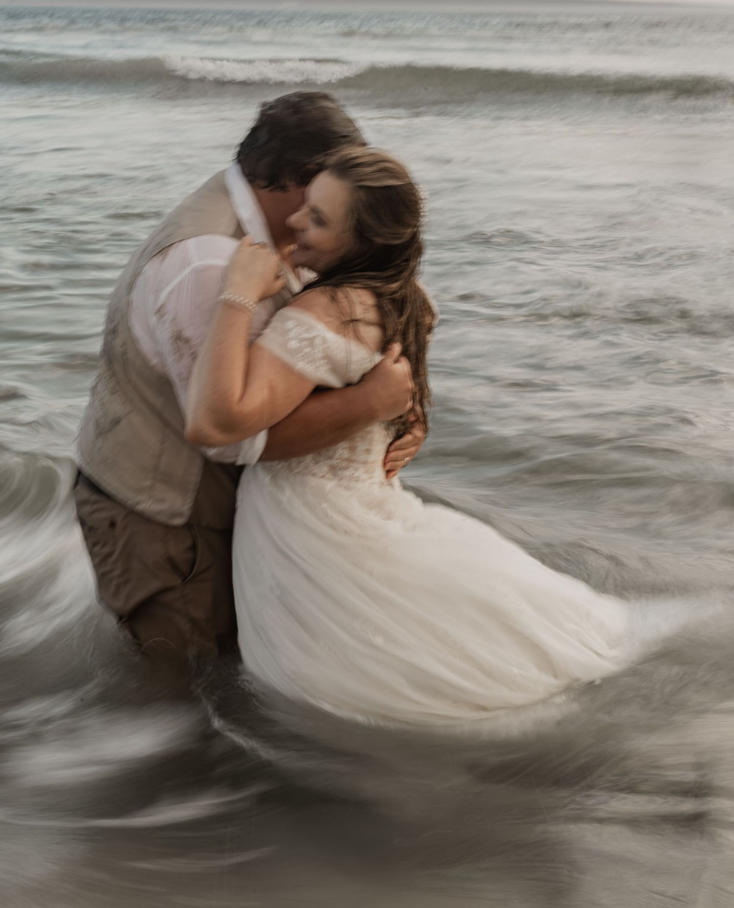 A couple embraces while standing in shallow ocean waves during a romantic beach photoshoot at sunset.