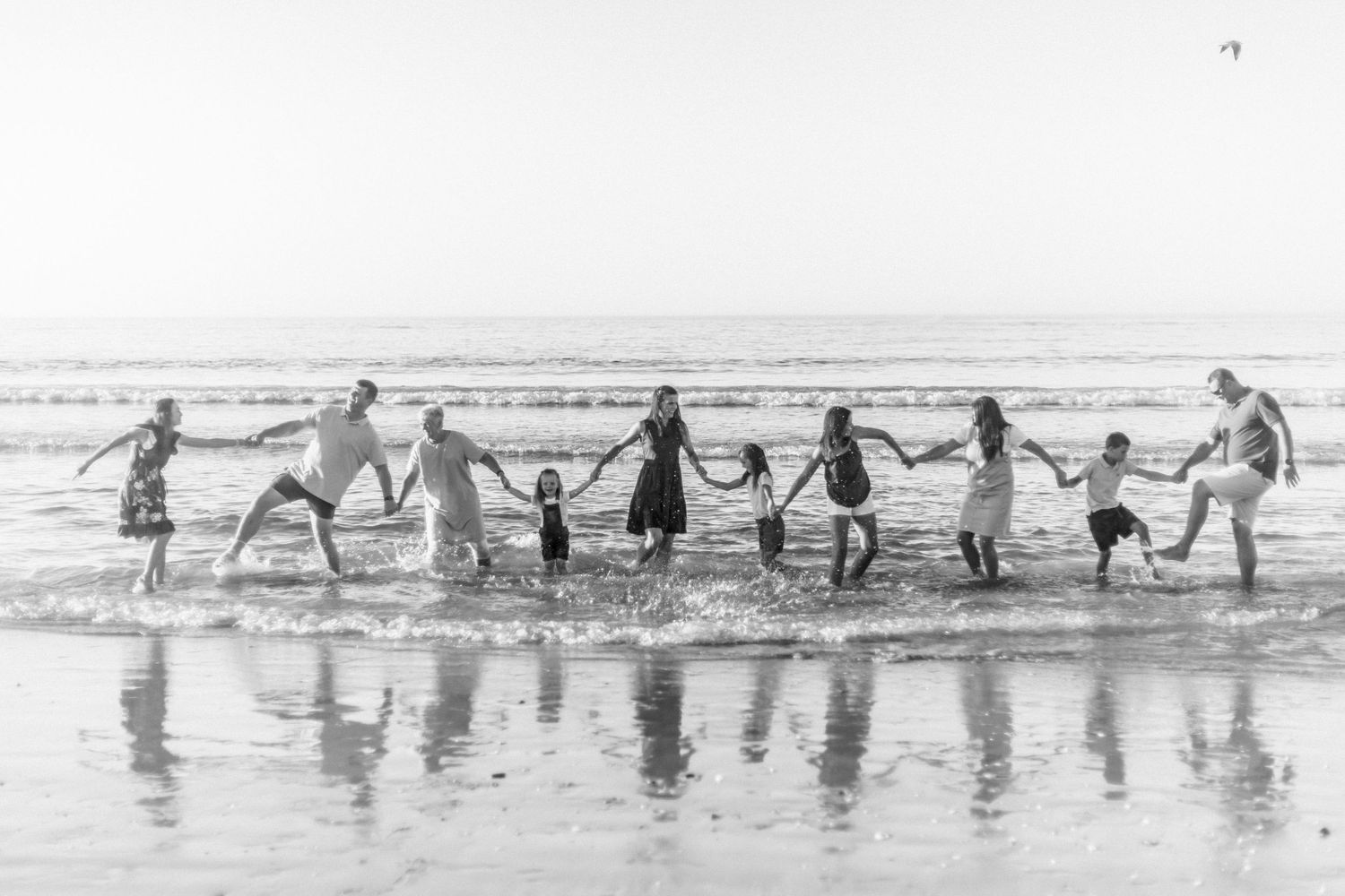 A group of people holding hands in a line at the beach, their reflections mirrored in the wet sand.