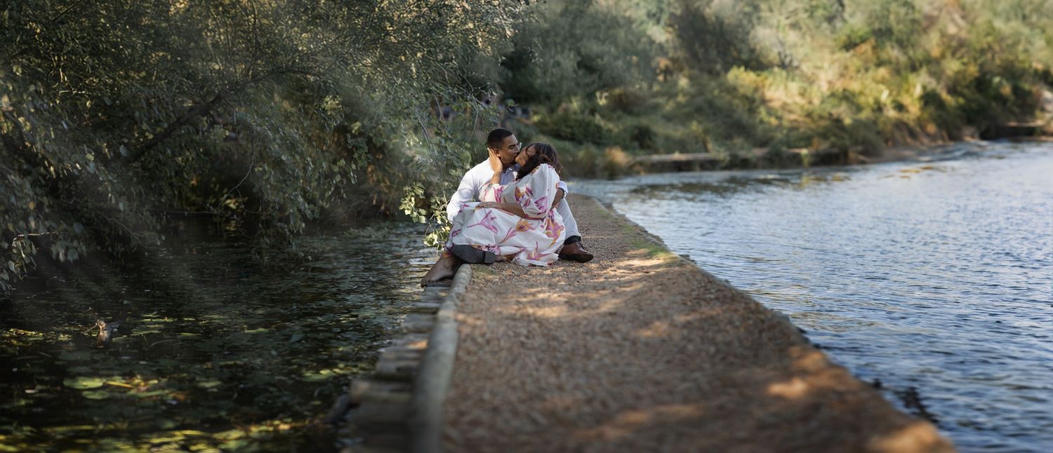 A person sits on a large rock by a peaceful river surrounded by lush green trees on a sunny day.