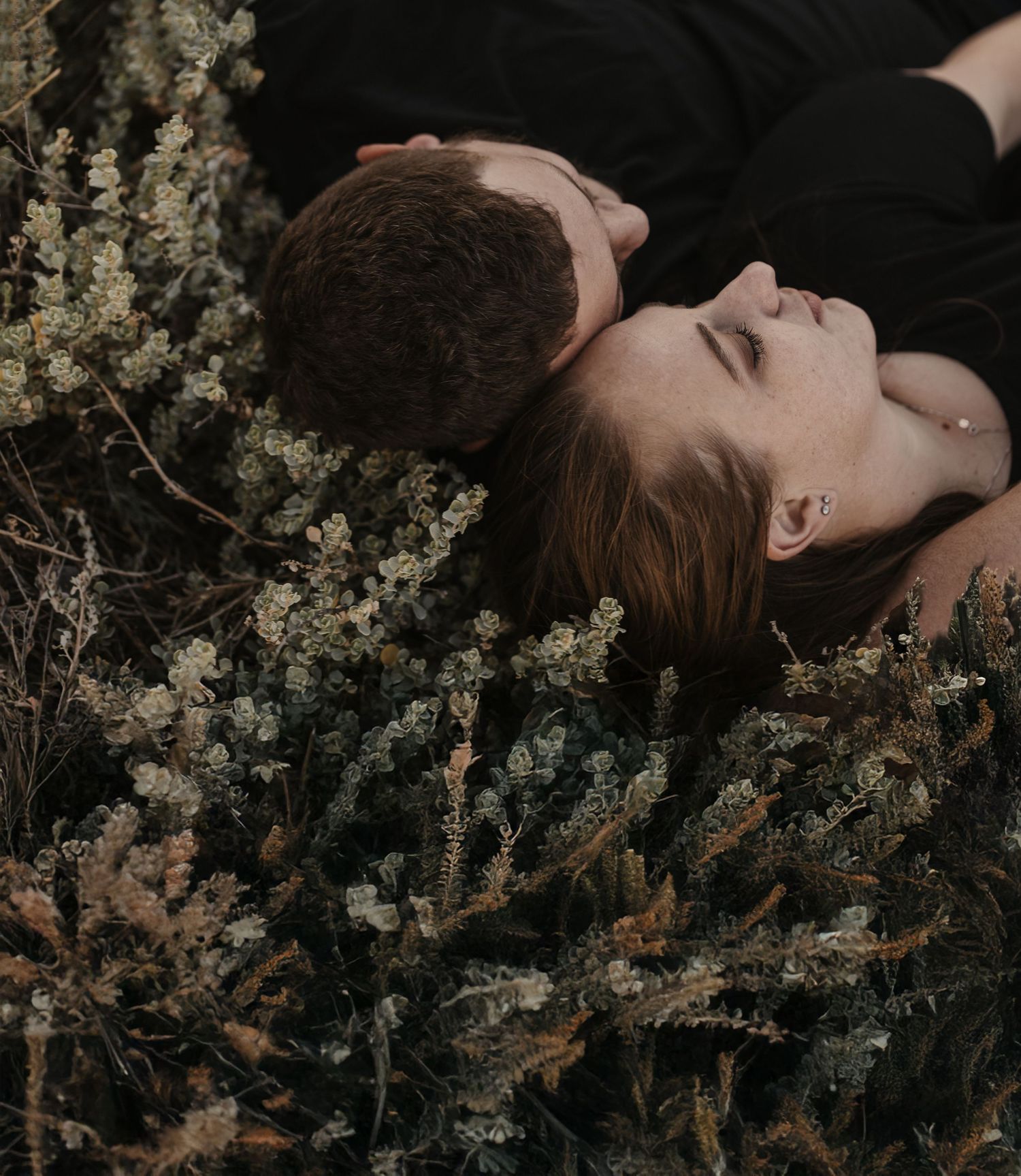 A couple lying in dark clothing among delicate white wildflowers in a moody, intimate outdoor setting.