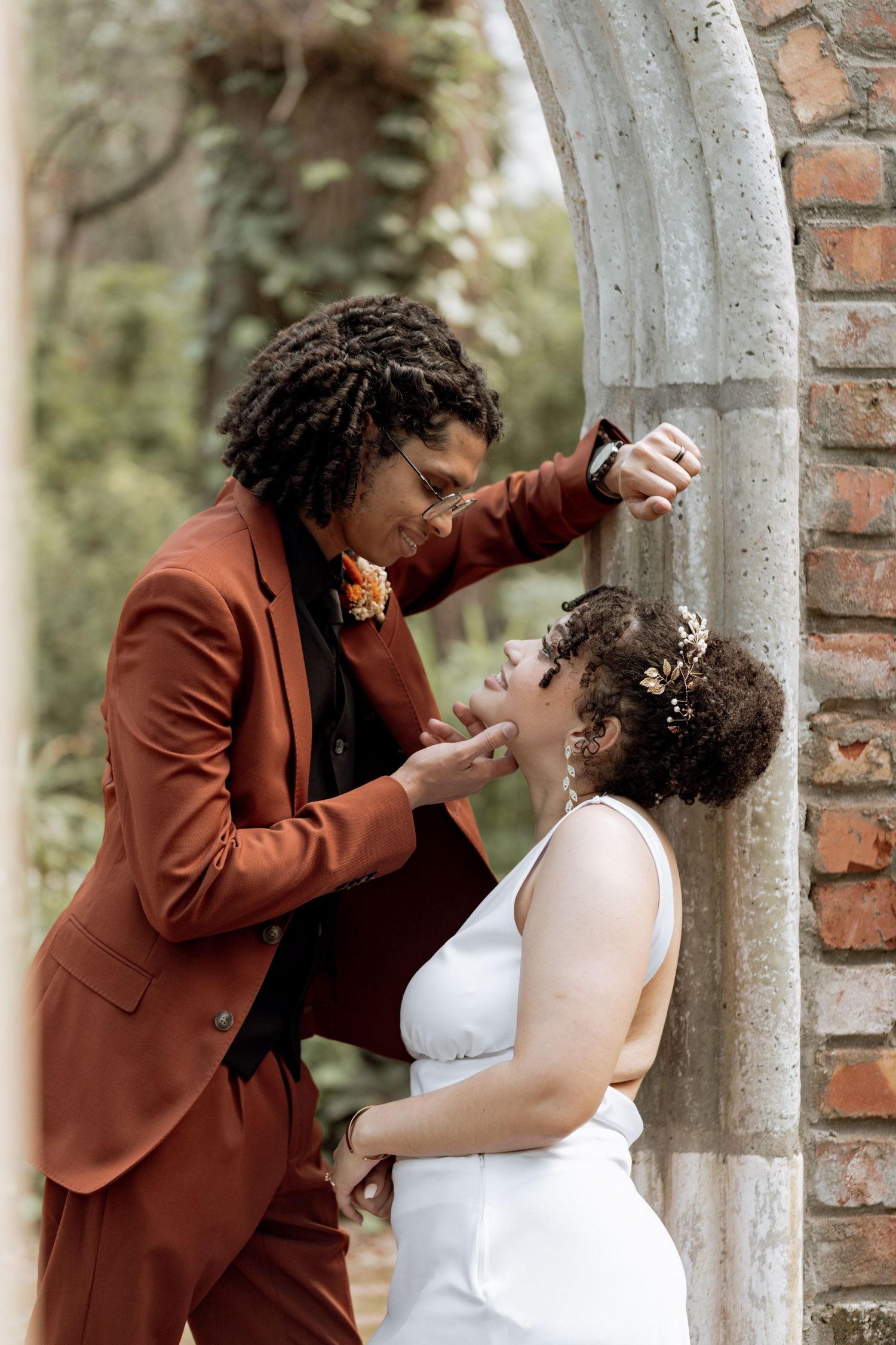 A couple shares a playful moment by a brick pillar while dressed in a burgundy suit and white dress.