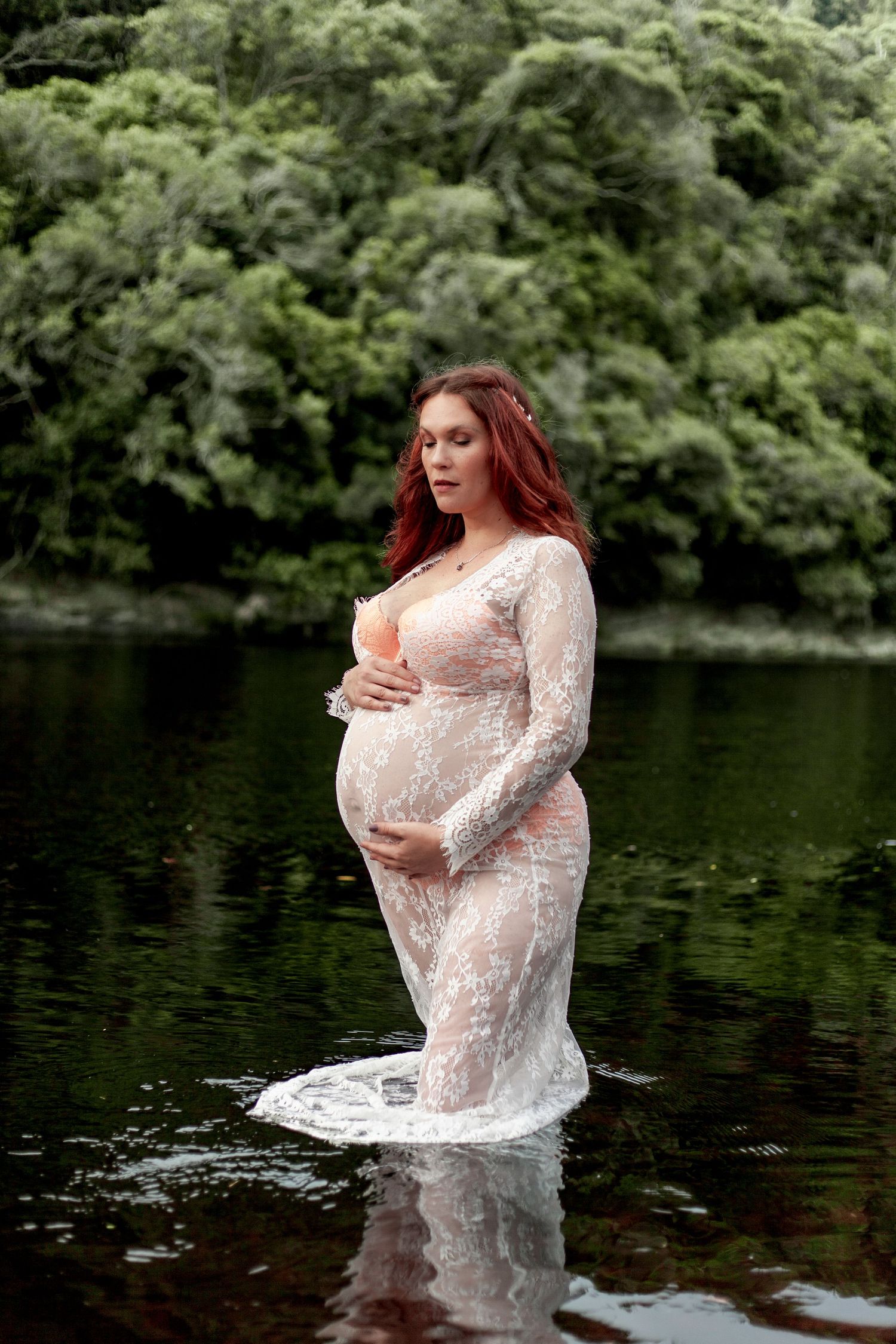 An ethereal maternity photoshoot in a tranquil lake shows a figure in a flowing white lace dress against lush greenery.