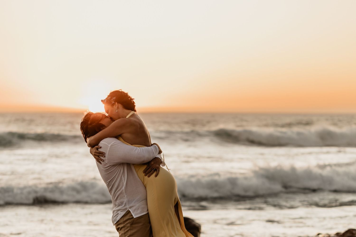 A couple embraces on a beach at sunset with crashing waves in the background creating a romantic silhouette.