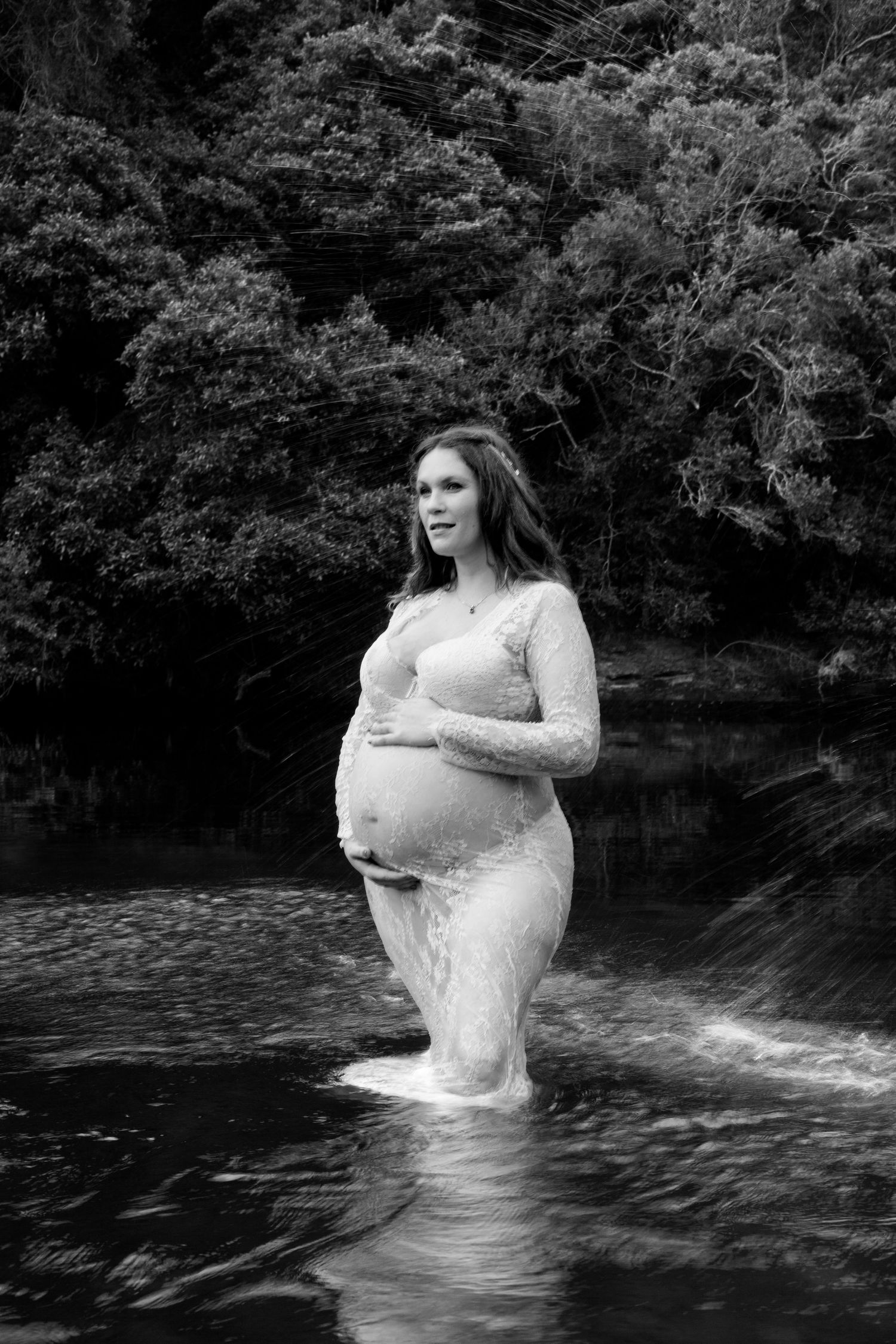 A moody black and white maternity portrait shows an expectant figure standing in flowing water surrounded by dense foliage.