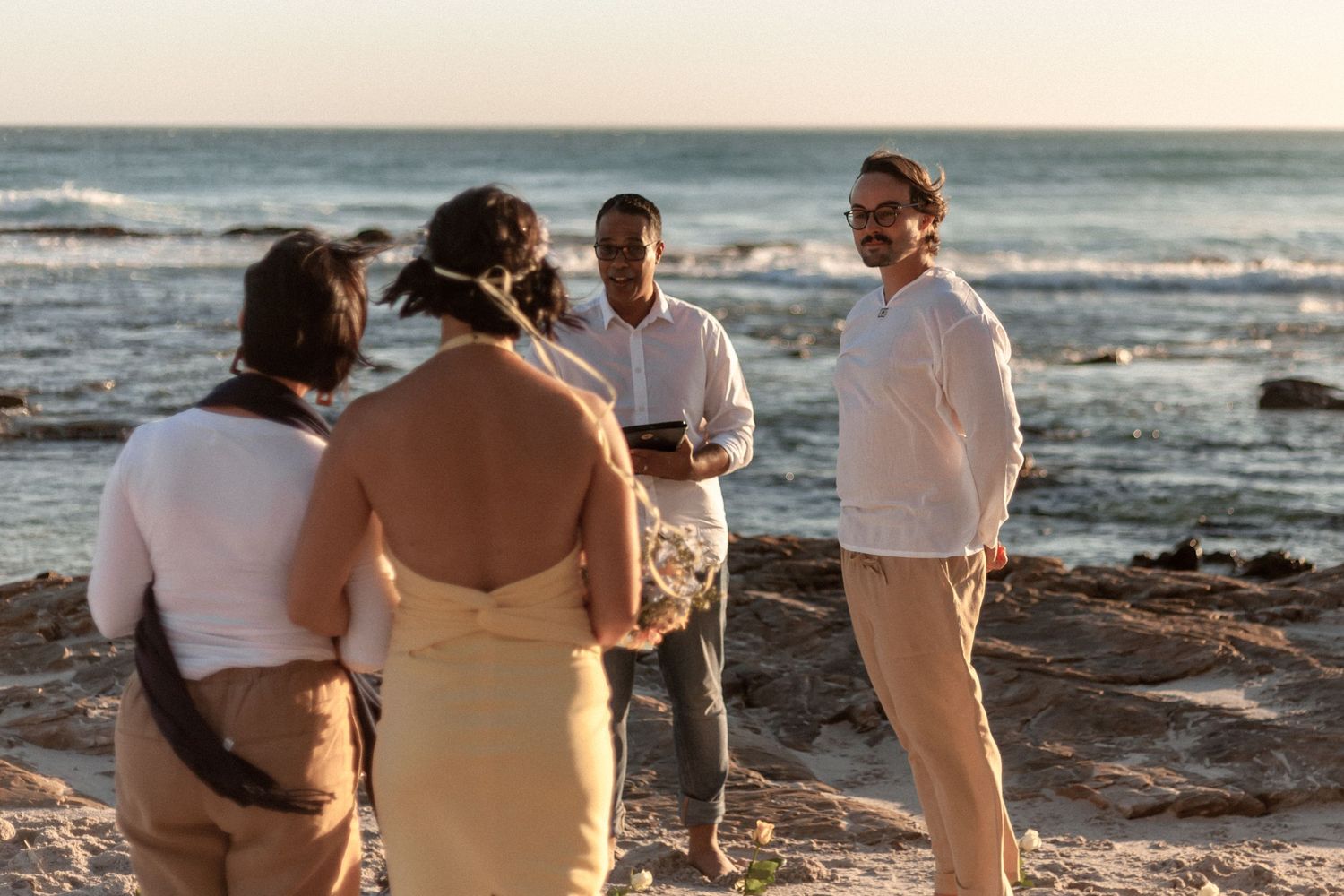 Wedding ceremony participants gather on a rocky beach at sunset with waves crashing in the background.