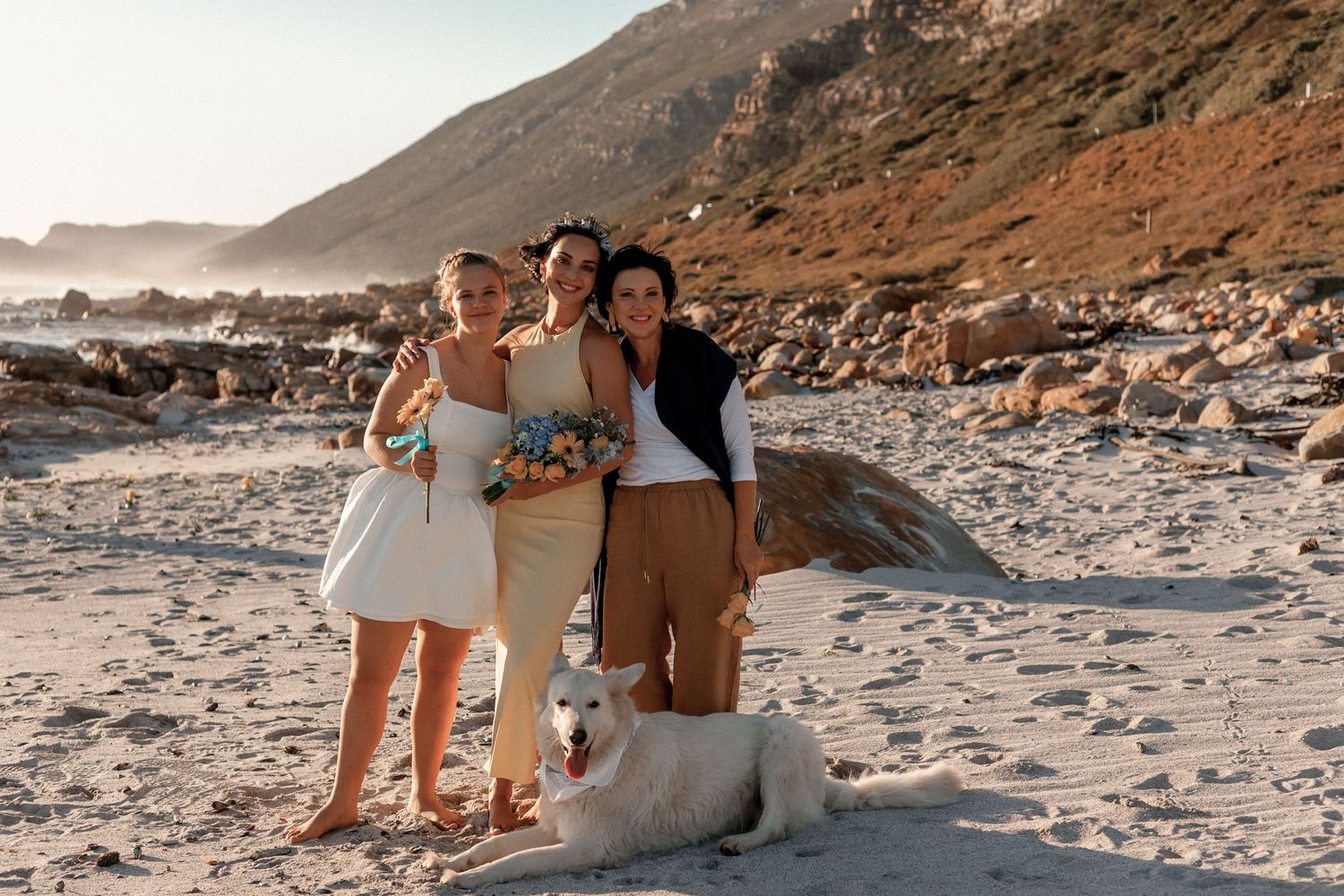 A group poses with their dog on a rocky beach at sunset with dramatic coastal cliffs in the background.