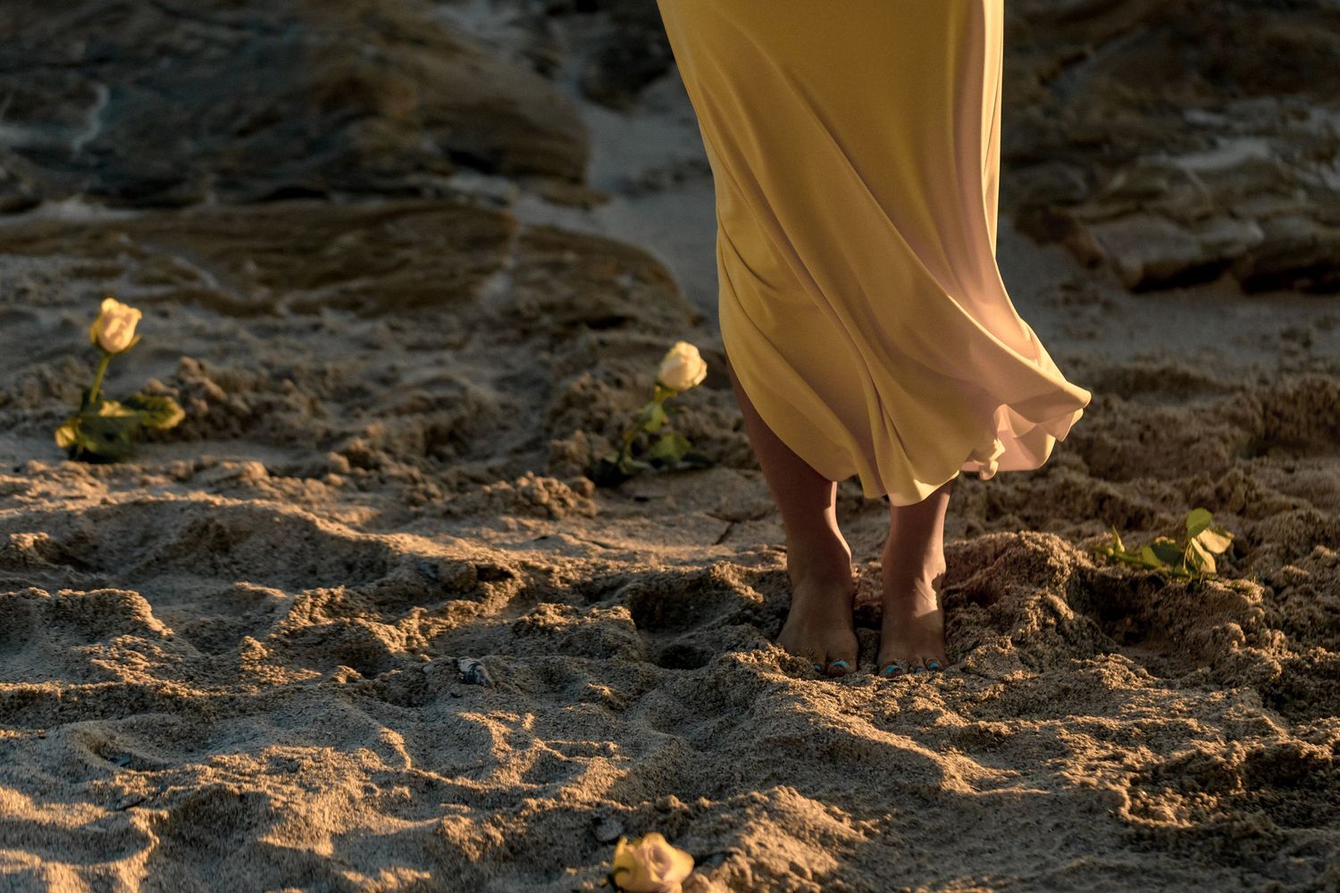 An artistic close-up view of bare feet walking on sand with a flowing dress at sunset.