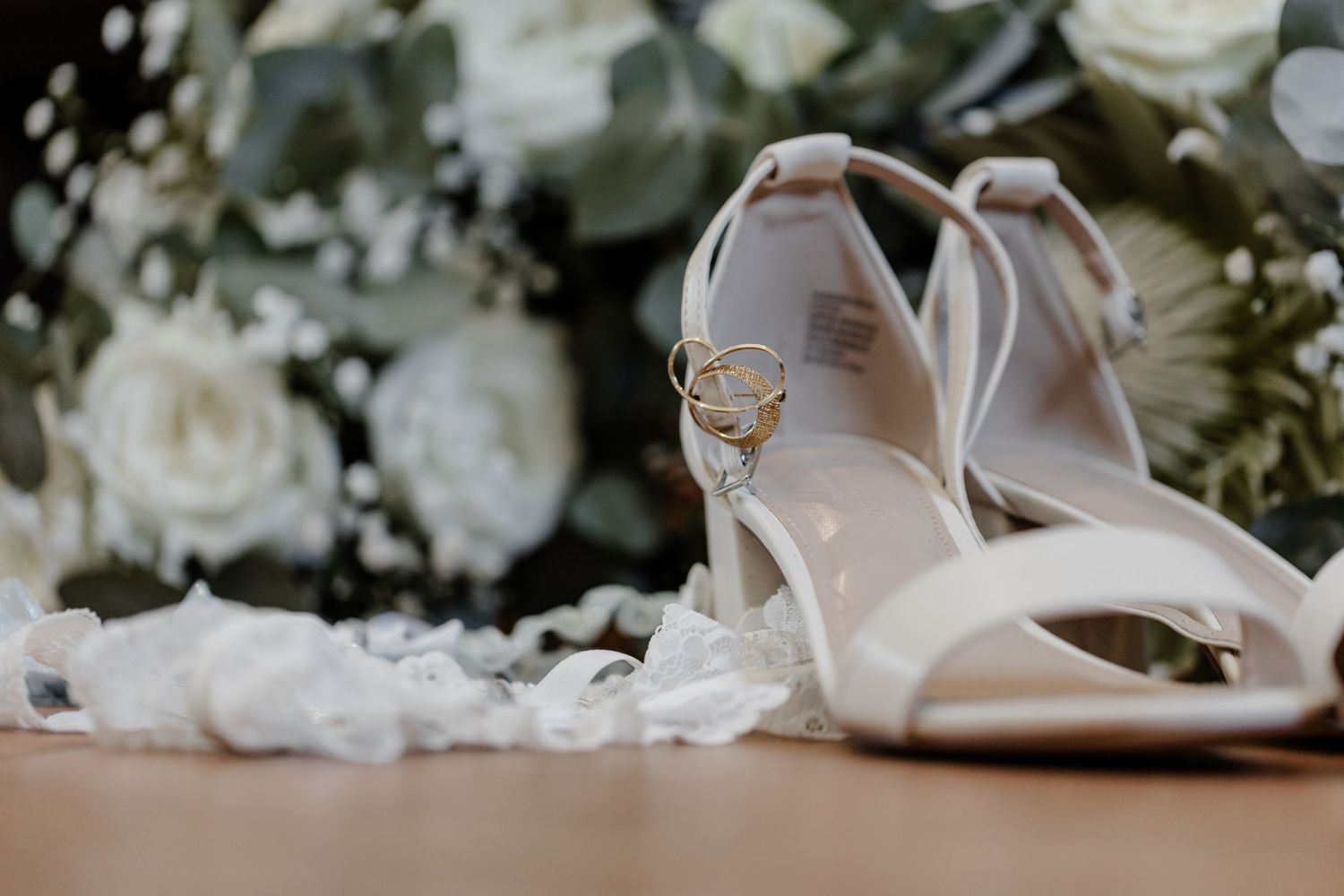 Elegant bridal shoes and wedding rings set against white floral arrangements and soft romantic lighting.