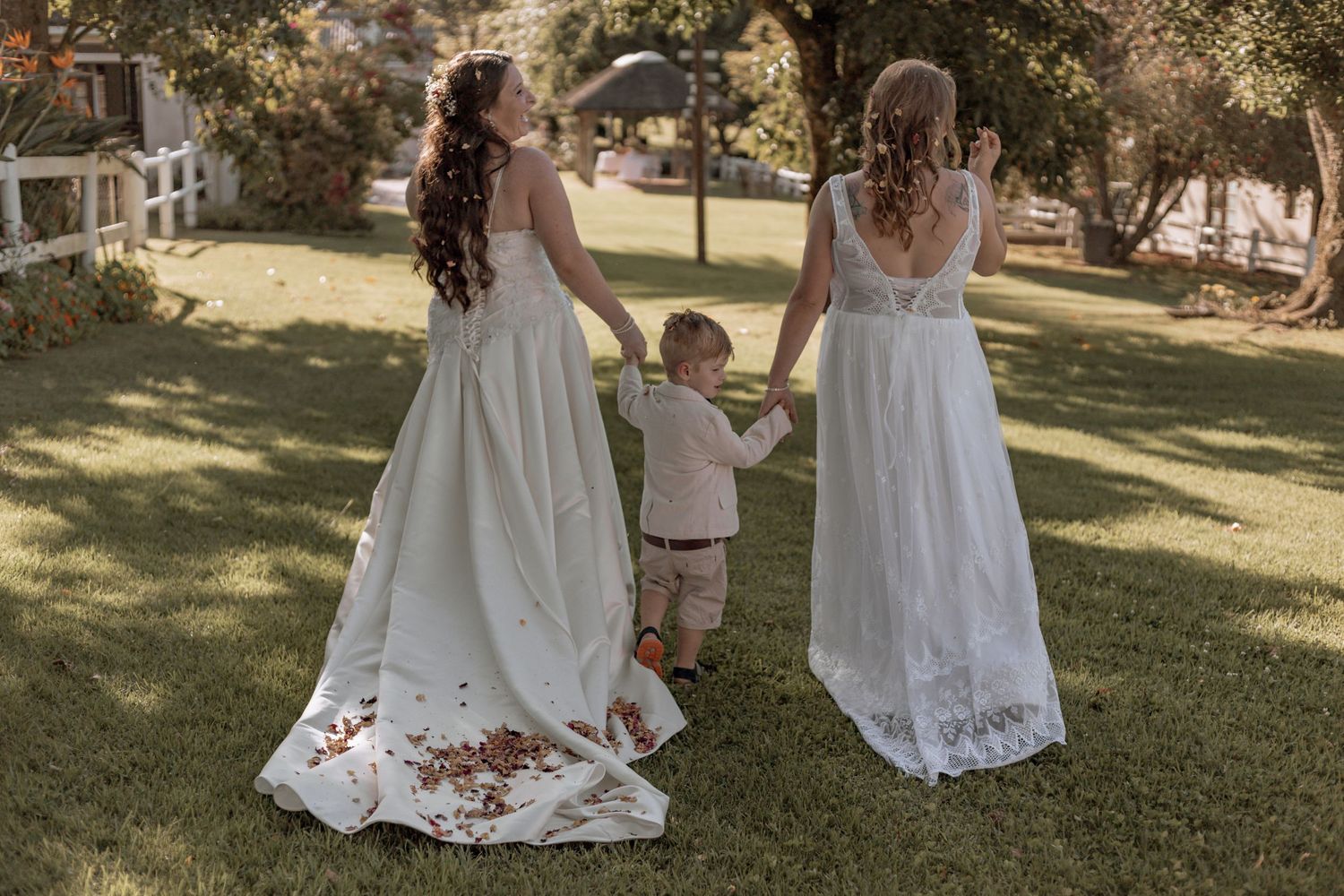 People in flowing white dresses pose together on a sunny lawn during what appears to be a wedding celebration.