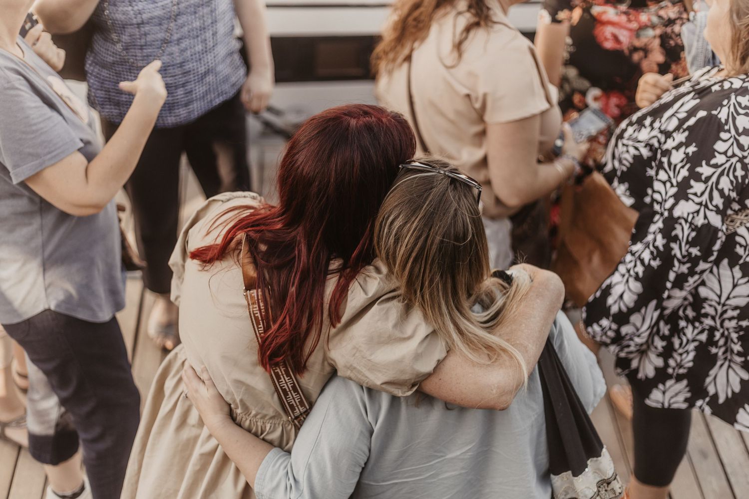 Three people embrace in a group hug wearing neutral toned clothing at a crowded gathering.