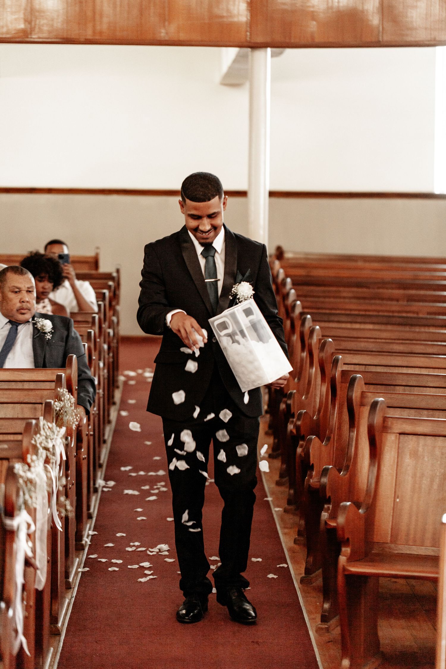 Ring bearers in formal black suits spread white petals down church aisle during wedding ceremony.