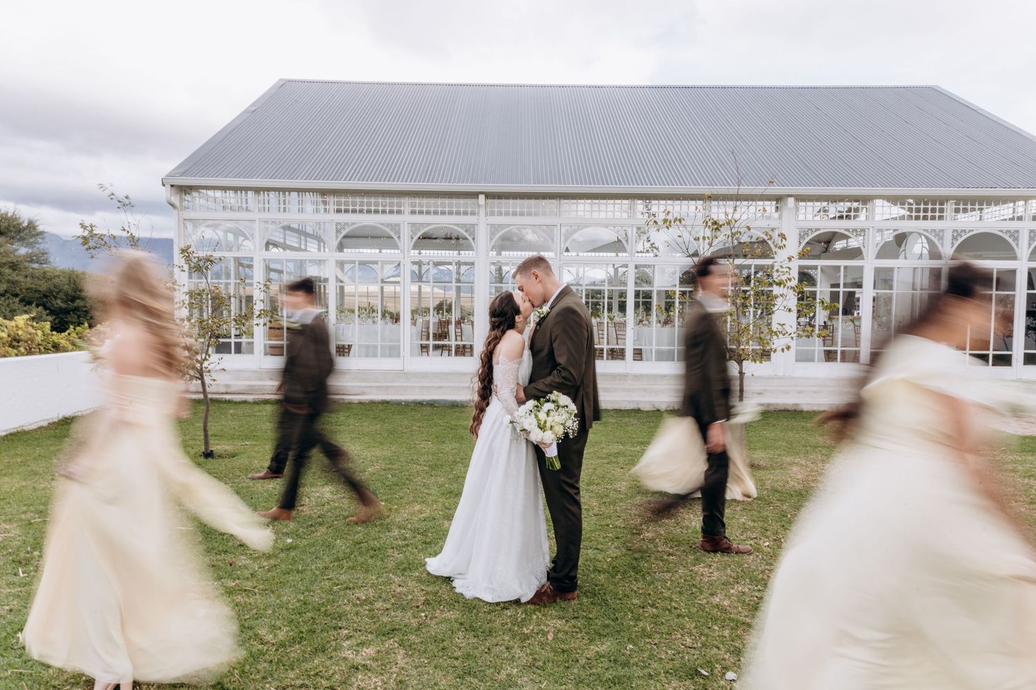 Bride and groom share a kiss outside a white greenhouse venue while wedding party walks past in soft motion blur.