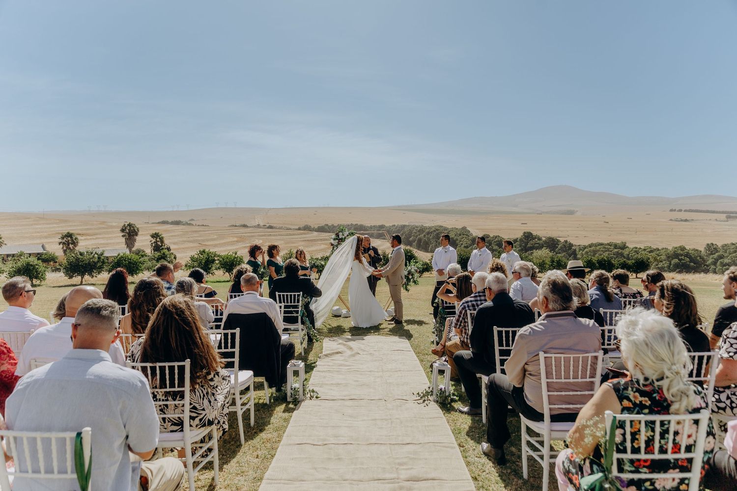 Outdoor wedding ceremony on a scenic hilltop with guests seated in white chairs facing the couple at the altar.