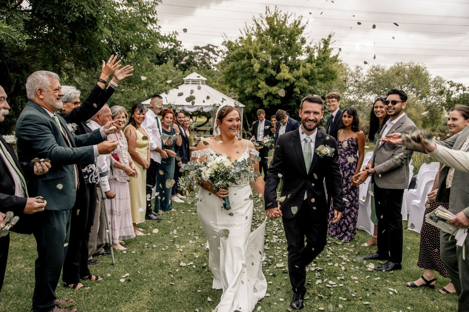Bride and groom walk through confetti shower surrounded by cheering guests at outdoor garden wedding ceremony.