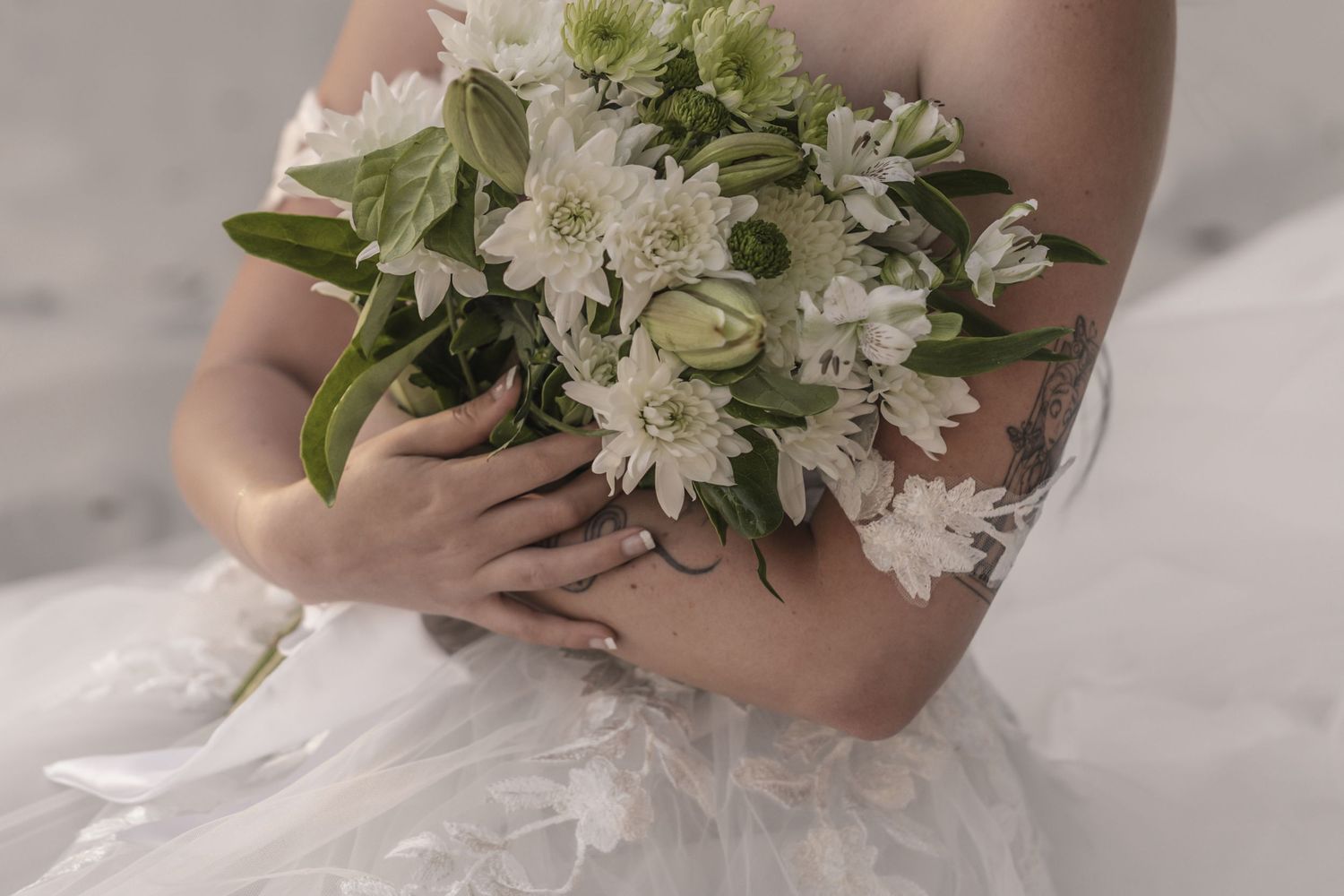 Bride in white lace gown holding a bouquet of white flowers, with a floral tattoo visible on her arm.