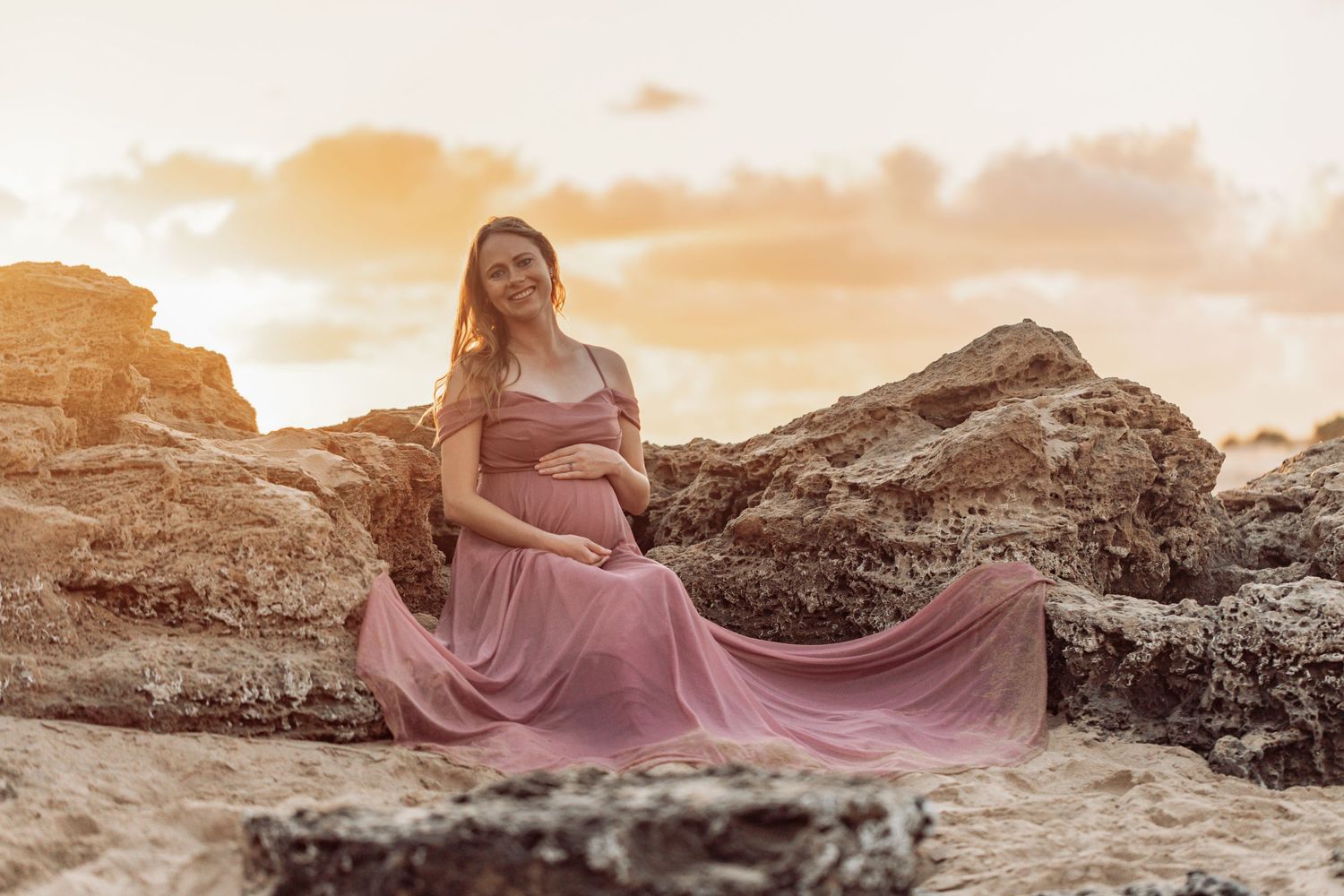 A flowing pink dress billows in the wind against a rocky coastal landscape at sunset.
