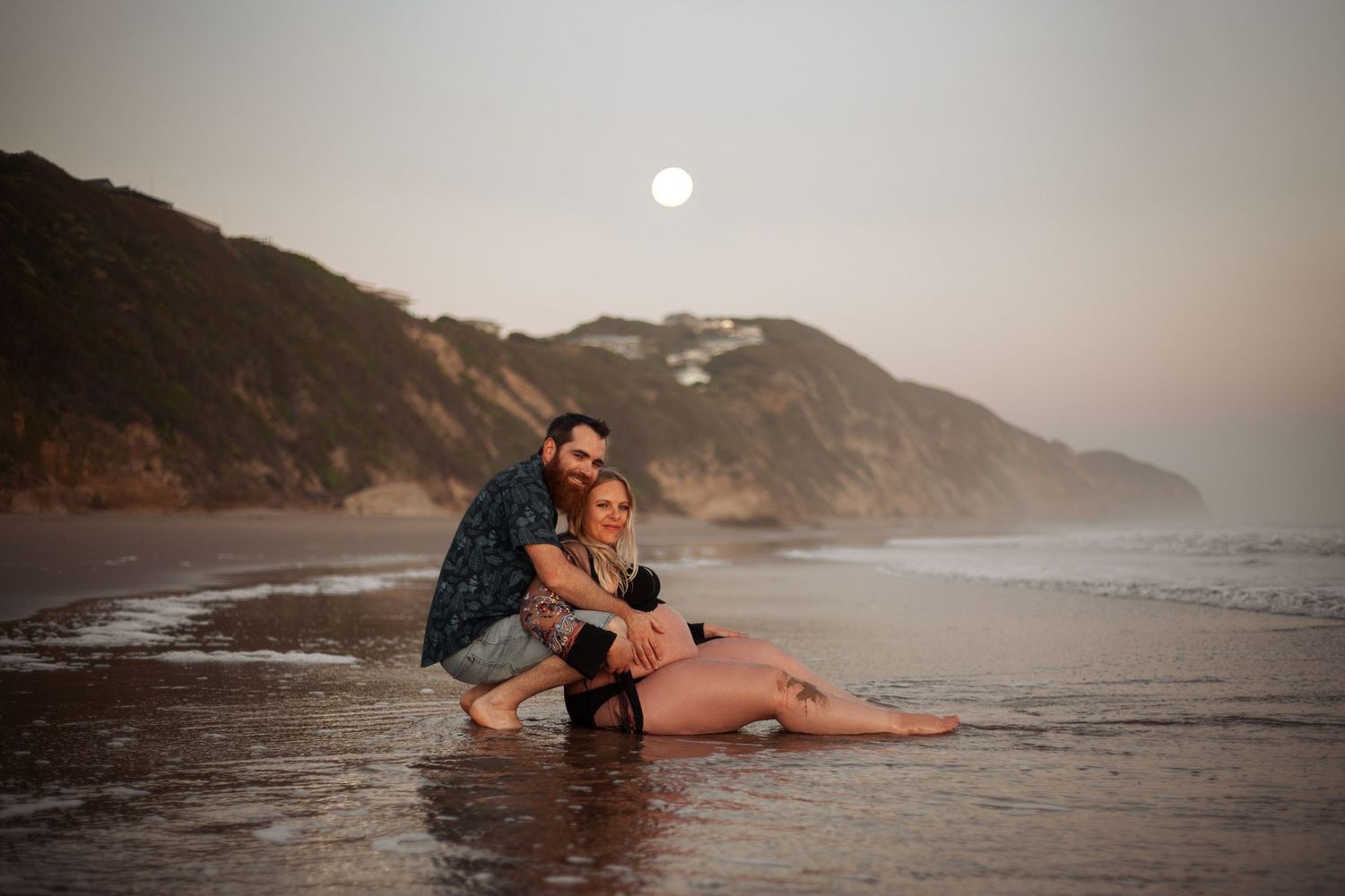 Two people embrace on a misty beach at sunset with rocky cliffs and ocean waves in the background.