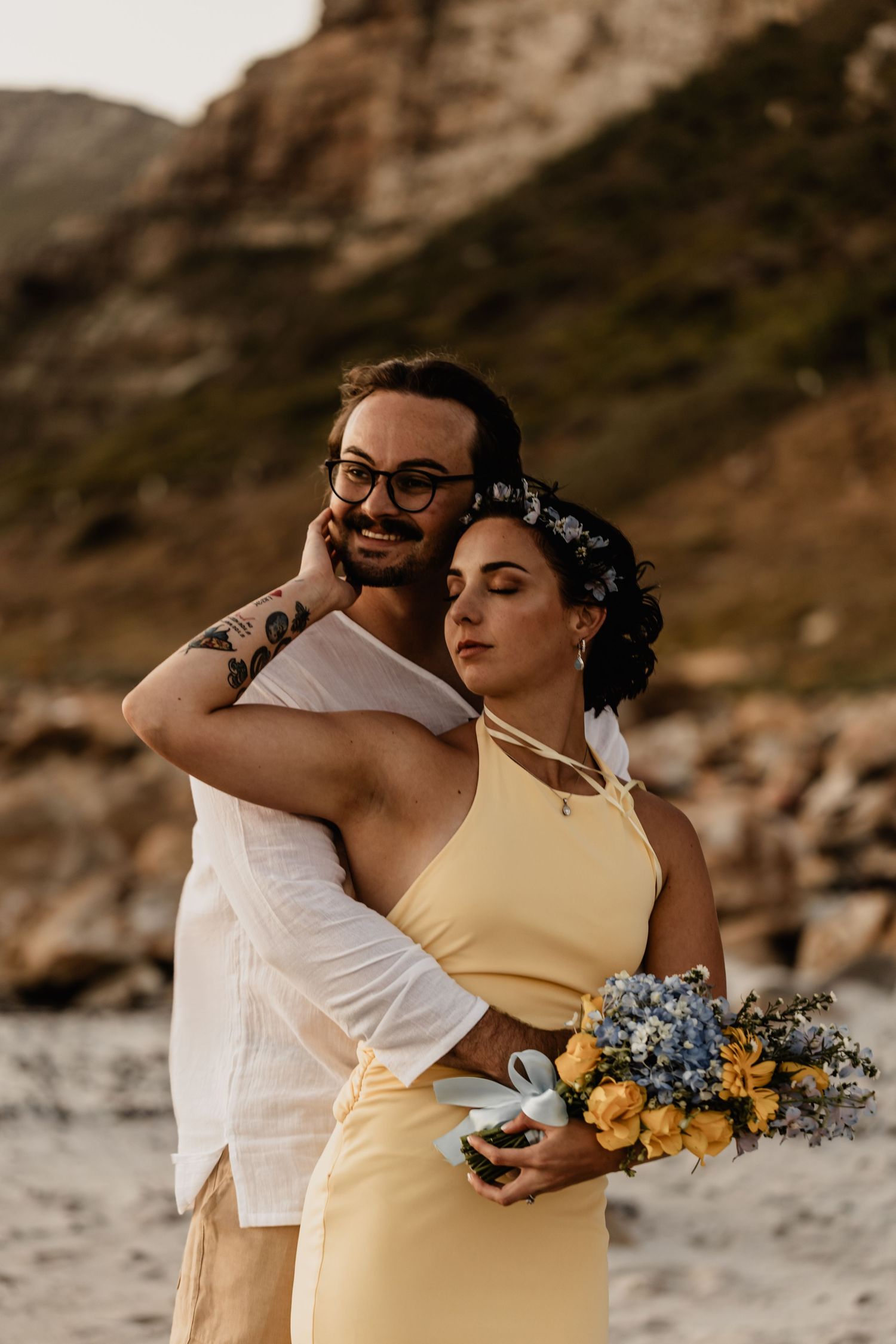 A couple embraces on a beach at sunset wearing yellow and white outfits with floral bouquet.