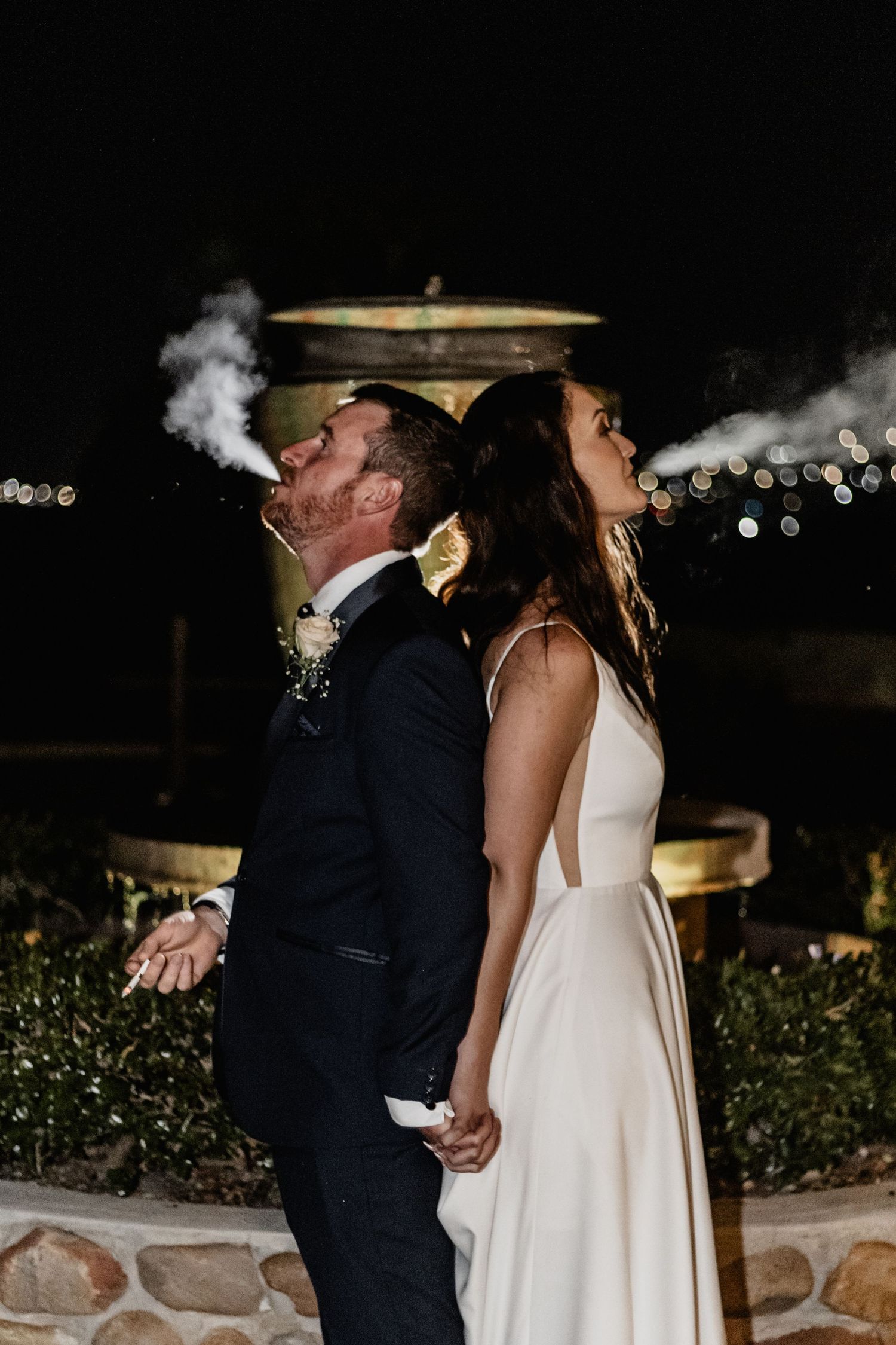A couple in wedding attire exhale smoke while standing together at night with bokeh lights in the background.