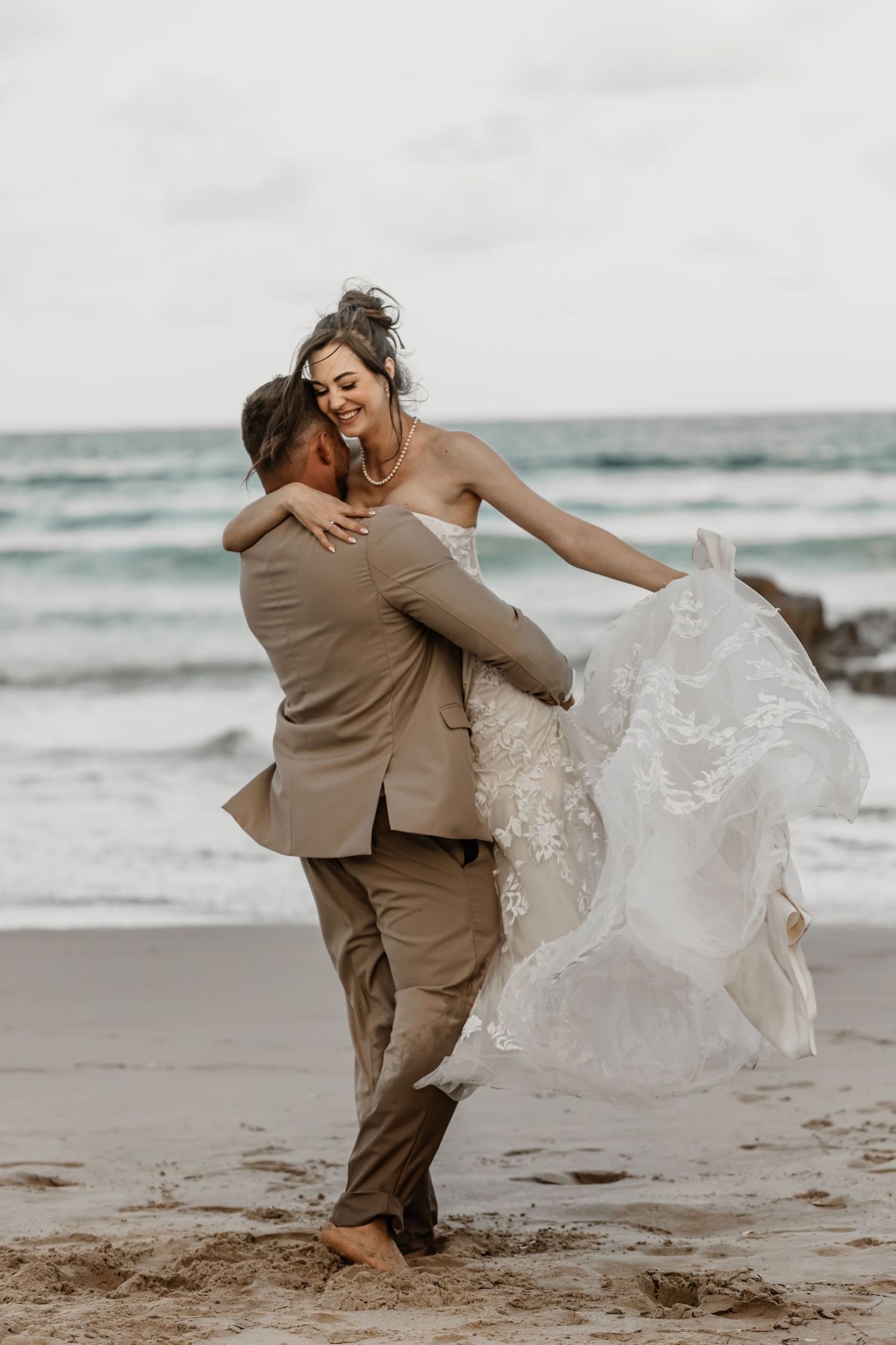 A joyful couple embraces on a beach, with the bride's white lace dress flowing in the ocean breeze.
