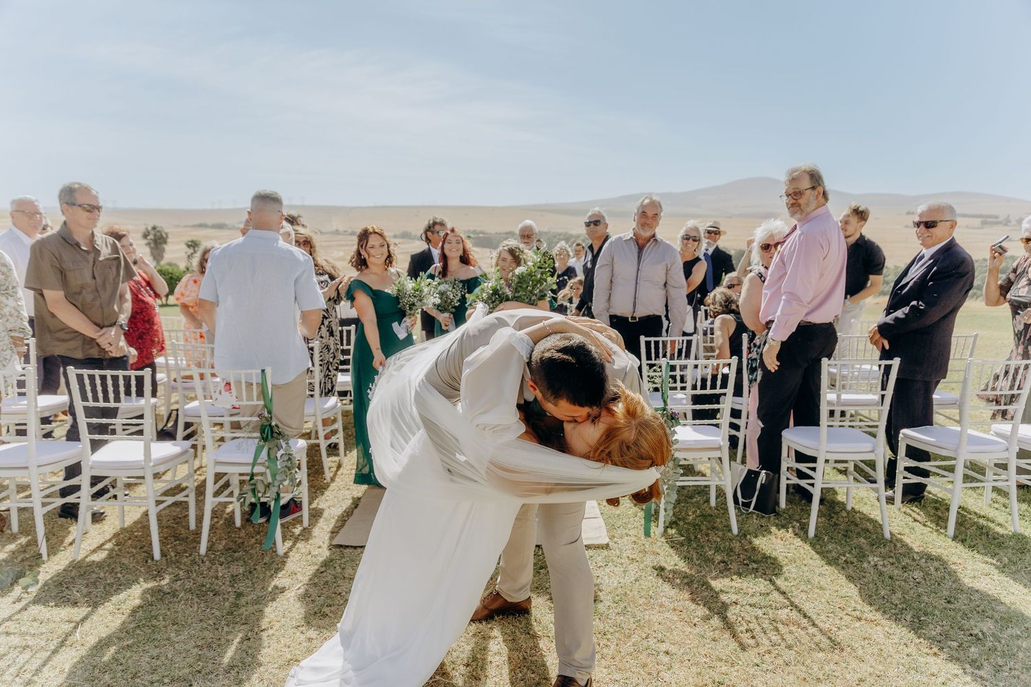 Bride and groom share a romantic dip kiss at outdoor desert wedding ceremony with white chairs and guests.