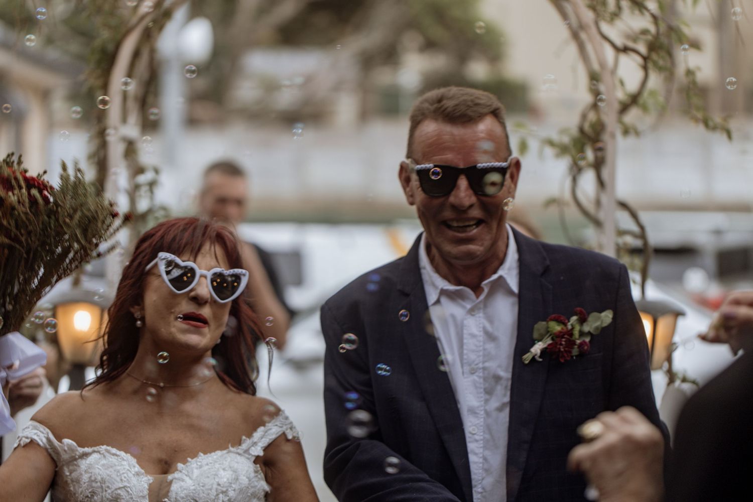 Joyful wedding party laughing together outdoors with flowers and string lights in background.