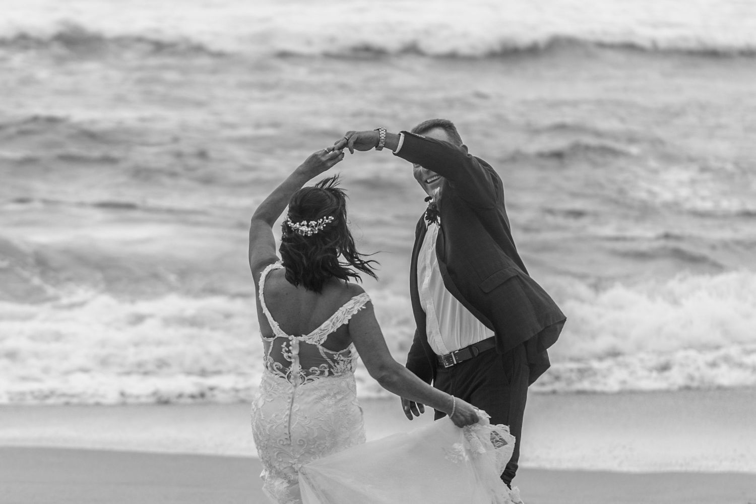 A couple dances together on a beach in black and white with ocean waves visible in the background.