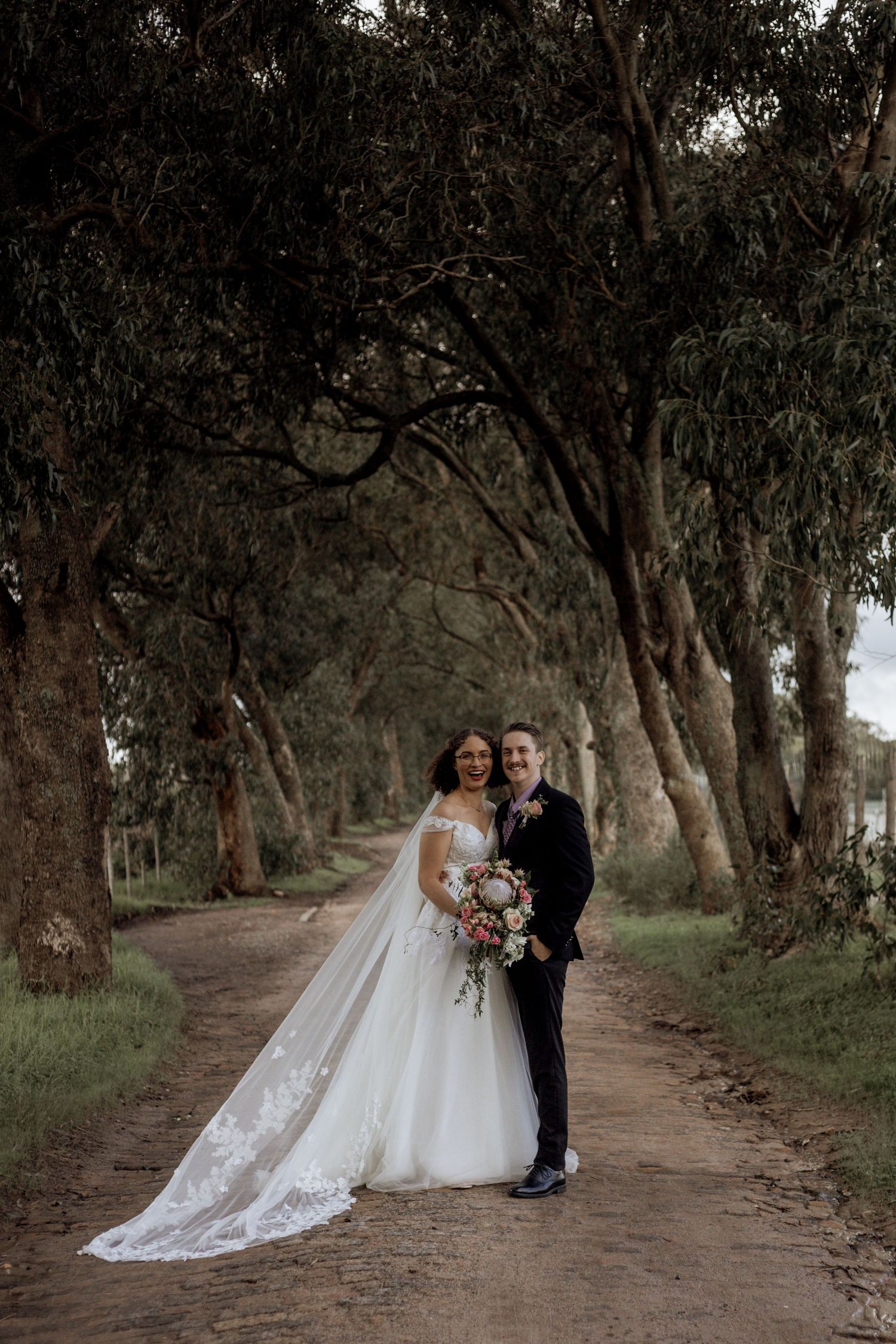 Newlyweds walk along a serene tree-lined dirt path during their rustic outdoor wedding photoshoot.