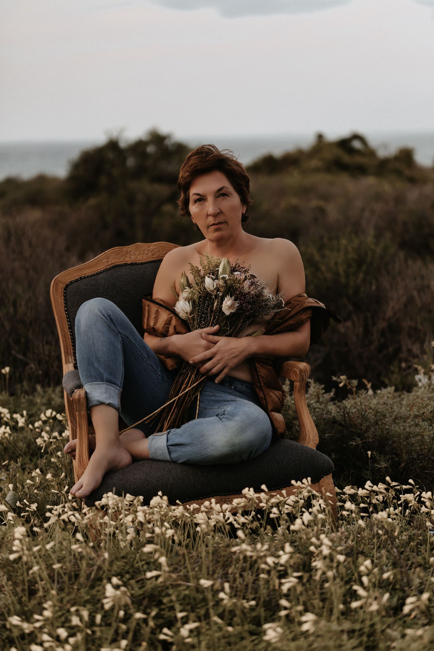 Moody portrait of someone sitting barefoot in vintage chair holding wildflowers amid a coastal meadow at sunset.