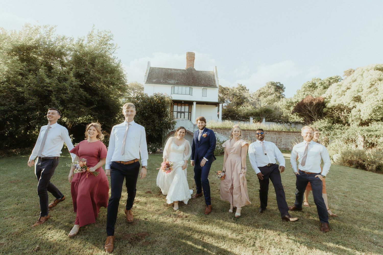 Wedding party poses together on a grassy lawn with a rustic house in the background on a sunny day.