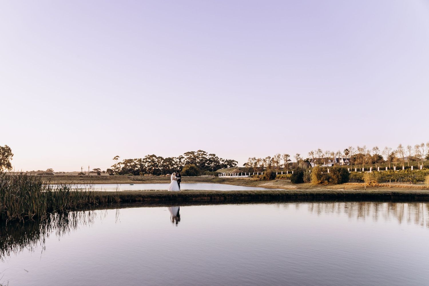 A couple stands by a tranquil reflective lake at dusk, surrounded by lush trees and reeds at a scenic wedding venue.